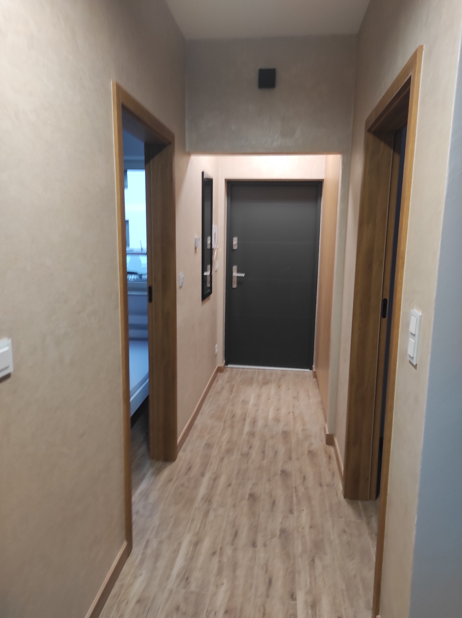 Korytarz po remoncie: drewnopodobne panele, beżowe ściany z efektem dekoracyjnym, drewniane ościeżnice drzwi i czarne drzwi wejściowe.