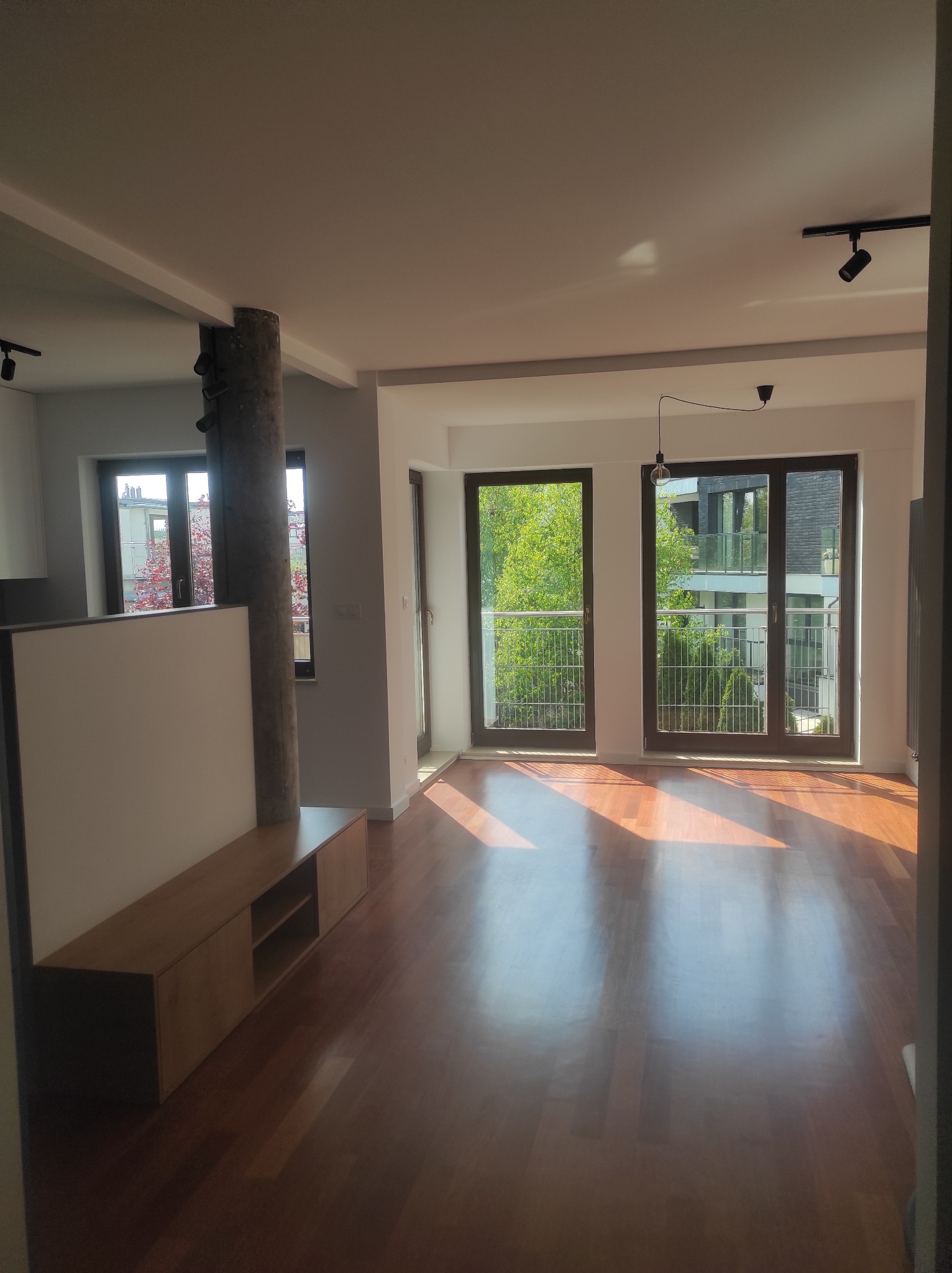 Jasne wnętrze z drewnianą podłogą i zabudową meblową. Widok na balkon przez duże okna. Elementy oświetlenia sufitowego i dekoracyjny filar.