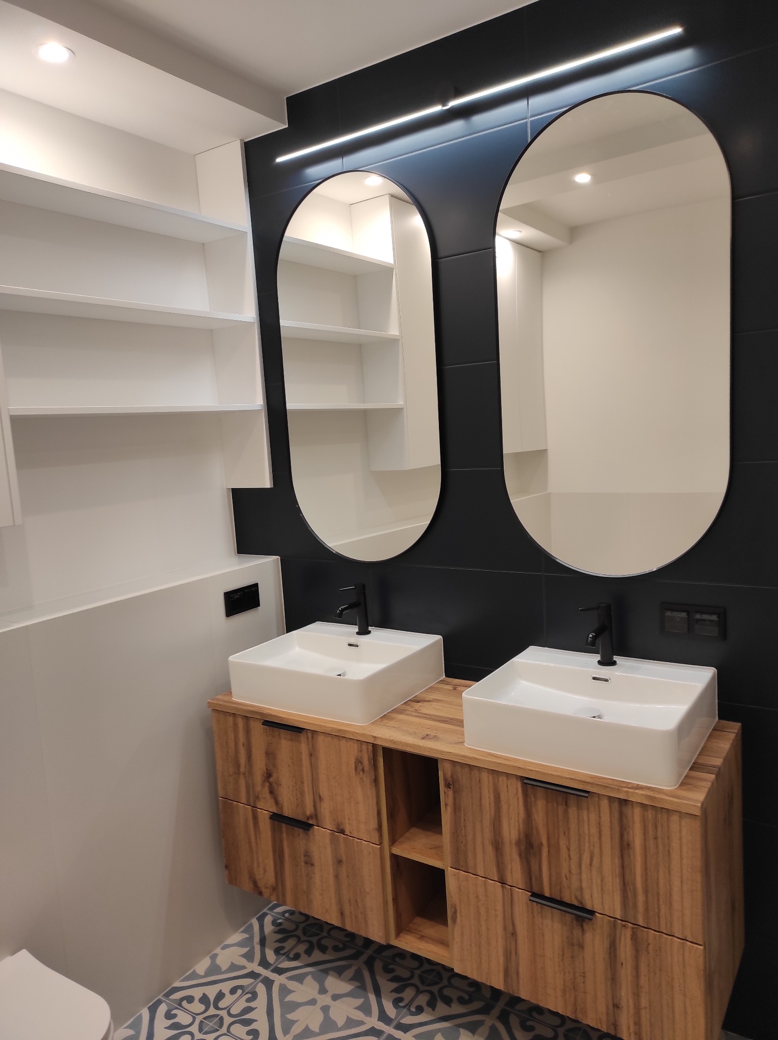 Nowoczesna łazienka z dwoma umywalkami nablatowymi na drewnianej szafce, czarne płytki i owalne lustra z podświetleniem LED. Białe półki w tle i wzorzysta podłoga.