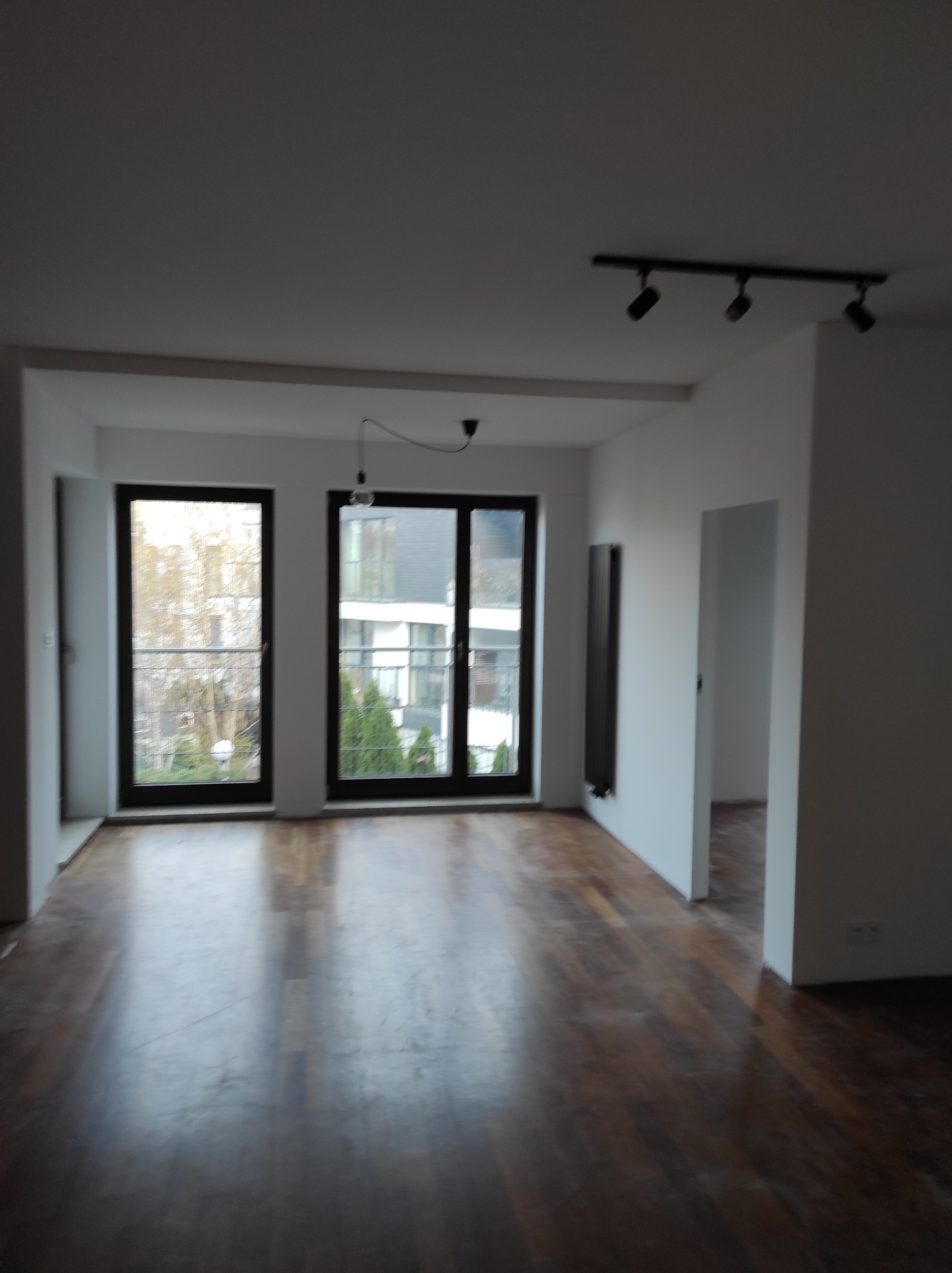 Puste, jasne wnętrze z parkietem, dużymi oknami i nowoczesnym oświetleniem punktowym. Widok na zieleń za oknem. Przygotowane do wykończenia.