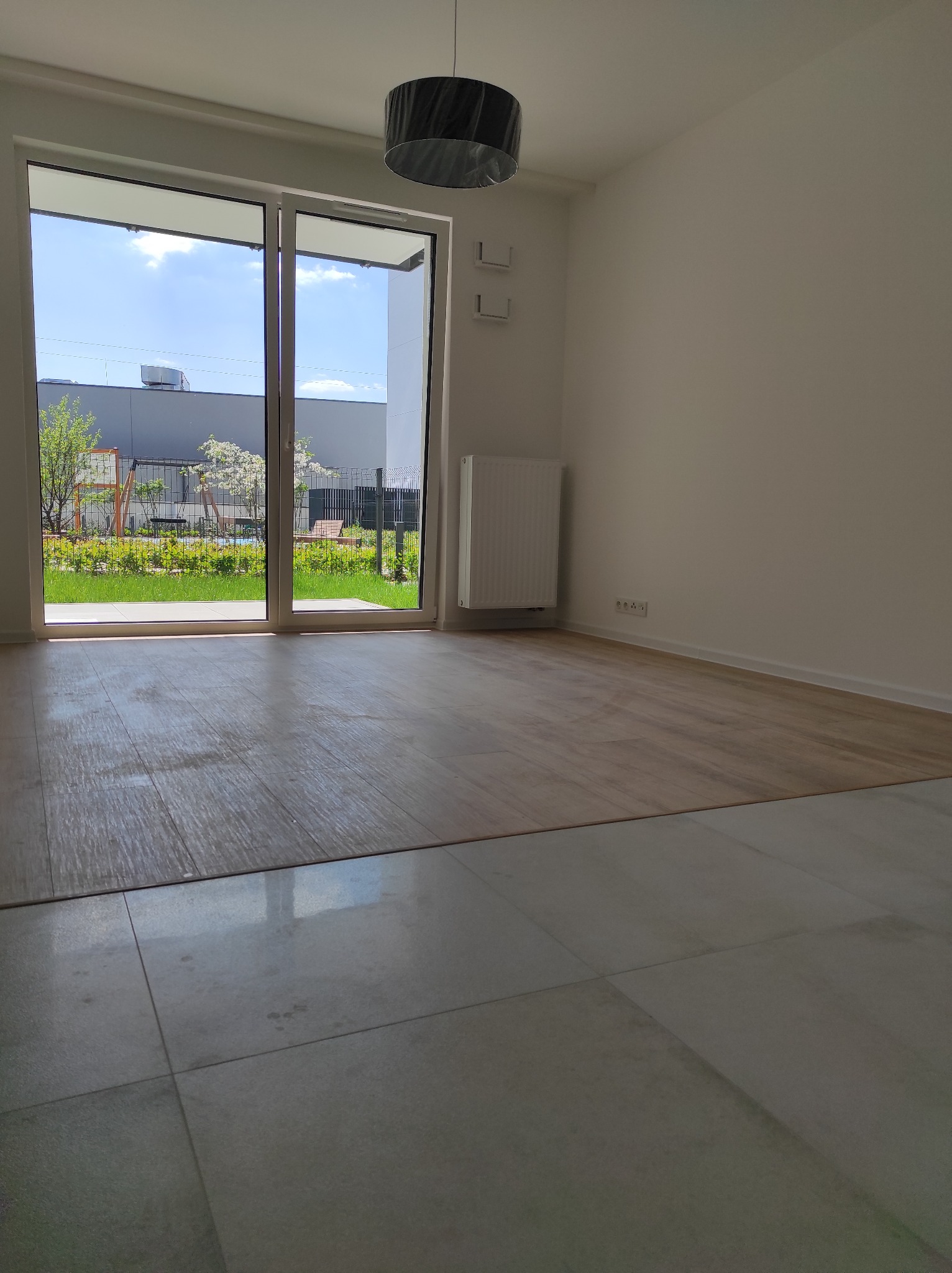 Jasne wnętrze z dużym oknem na ogród. Częściowo ułożona podłoga z paneli przechodzi w płytki. Widoczny grzejnik i lampa sufitowa.