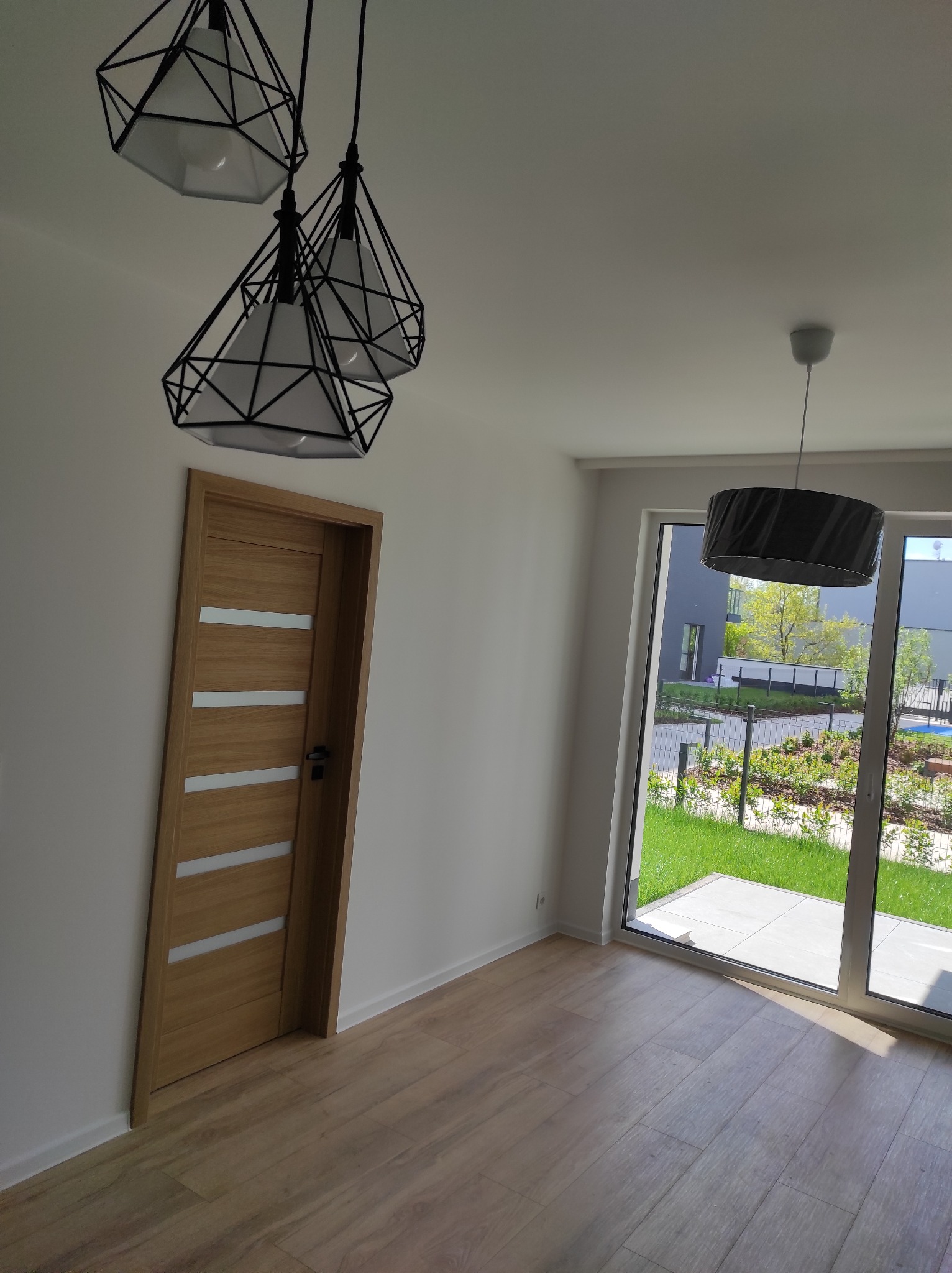 Jasne wnętrze z drewnianą podłogą, nowoczesnymi lampami i drzwiami z przeszkleniami. Widok na zieleń za oknem. Minimalistyczny design.