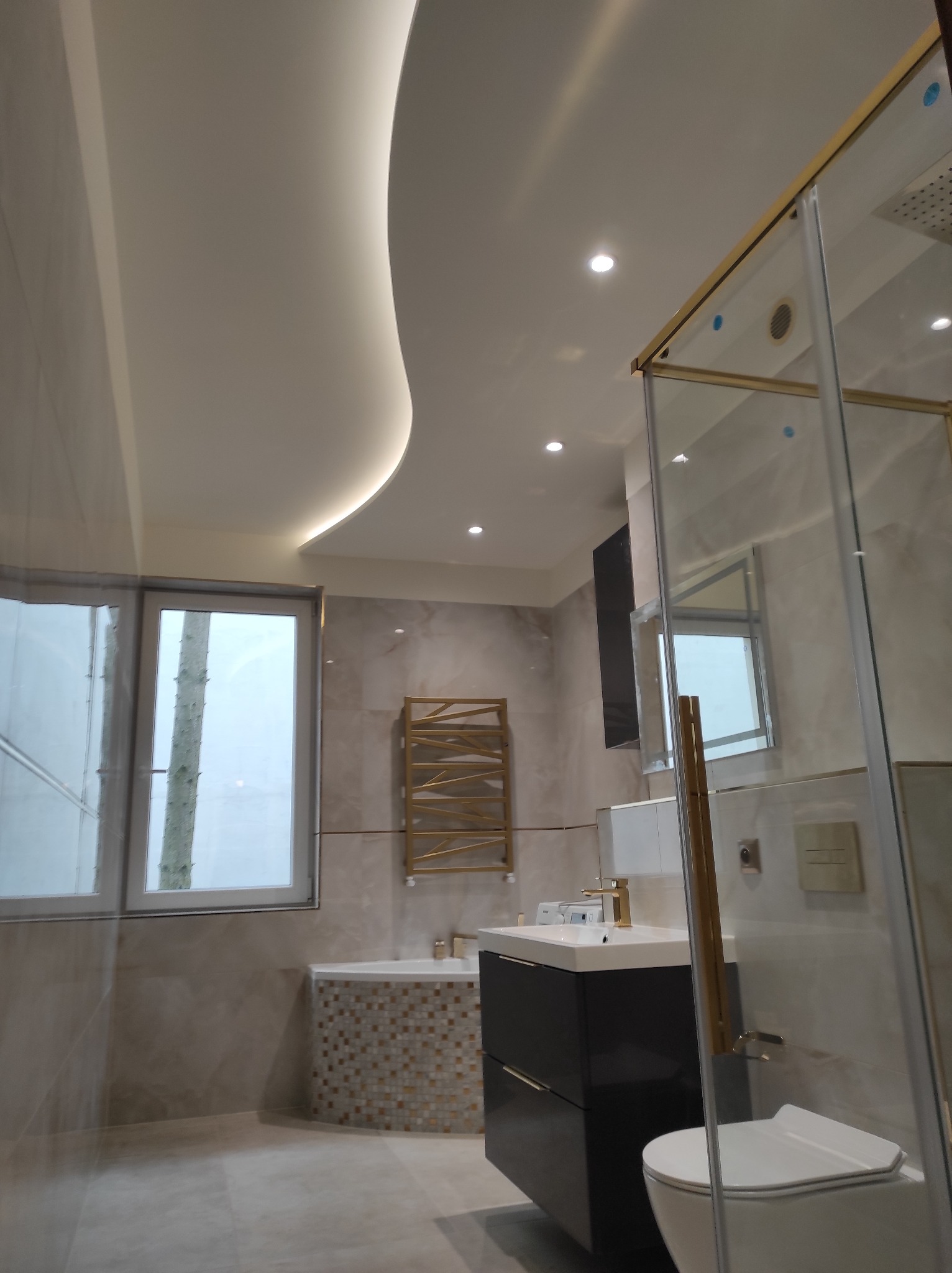 Nowoczesna łazienka z kabiną prysznicową w złotych ramach, umywalką z szafką i wanną obłożoną mozaiką. Sufit podwieszany z oświetleniem LED.