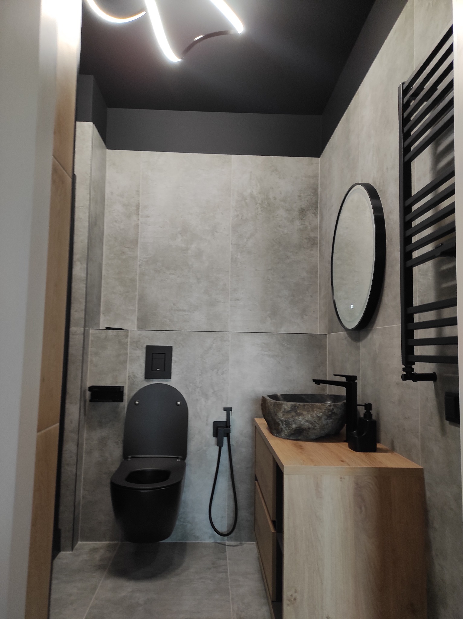 Nowoczesna łazienka z szarymi płytkami, czarną armaturą i umywalką z kamienia. Oświetlenie sufitowe LED dodaje elegancji. Minimalistyczny design.