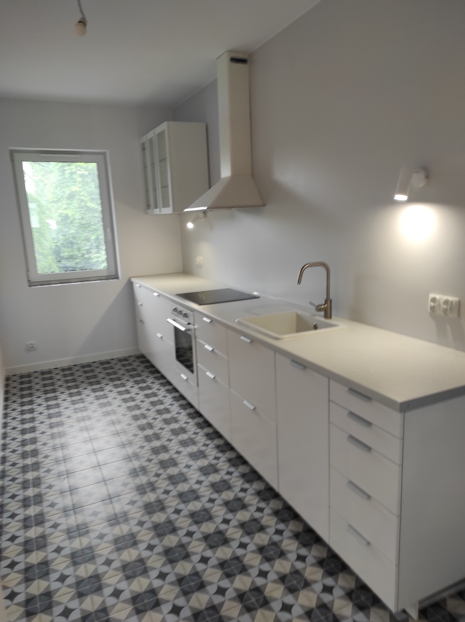 Jasna kuchnia z białymi meblami i wzorzystą podłogą. Minimalistyczny design, okno z widokiem na zieleń. Nowoczesne oświetlenie punktowe.