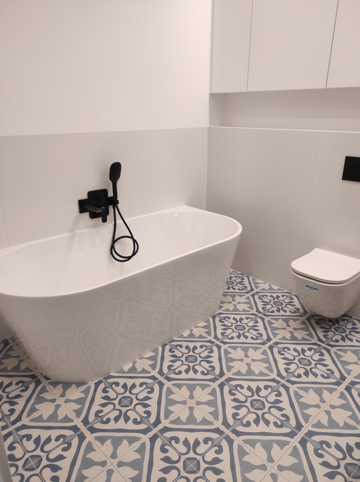 Biała wanna wolnostojąca z czarną armaturą i wisząca toaleta na podłodze w niebiesko-białe wzory. Minimalistyczna łazienka z białymi szafkami.