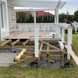 SKANDWOOD - Budowa tarasu drewnianego z białą pergolą. Widoczna konstrukcja nośna, częściowo ułożona deska tarasowa i pracownik montujący balustradę. W tle dom jednorodzinny.