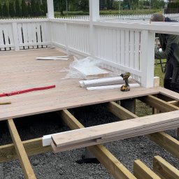 SKANDWOOD - Budowa tarasu: drewniana konstrukcja szkieletowa z częściowo ułożonymi deskami, biała balustrada i pergola w tle. Widoczne narzędzia i materiały budowlane.