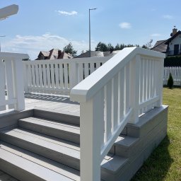 SKANDWOOD - Jasnoszary taras z białymi balustradami i schodami, widok z dołu, w tle domy i błękitne niebo. Estetyczne wykończenie, schludny design.