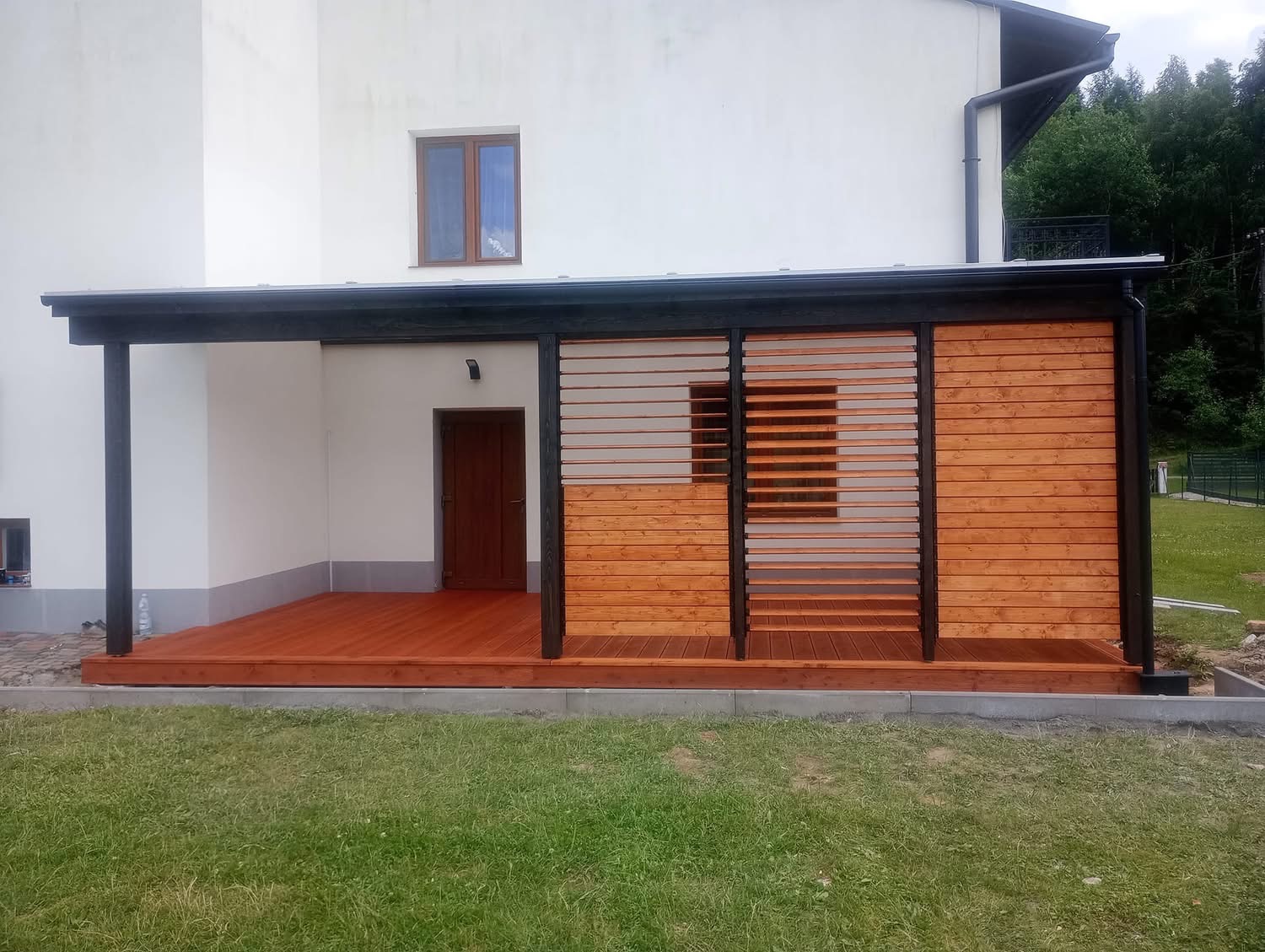 Zadaszony taras z drewnianą podłogą i ażurową ścianą z desek, przylegający do białego budynku. Ciemne, kontrastowe elementy konstrukcyjne.