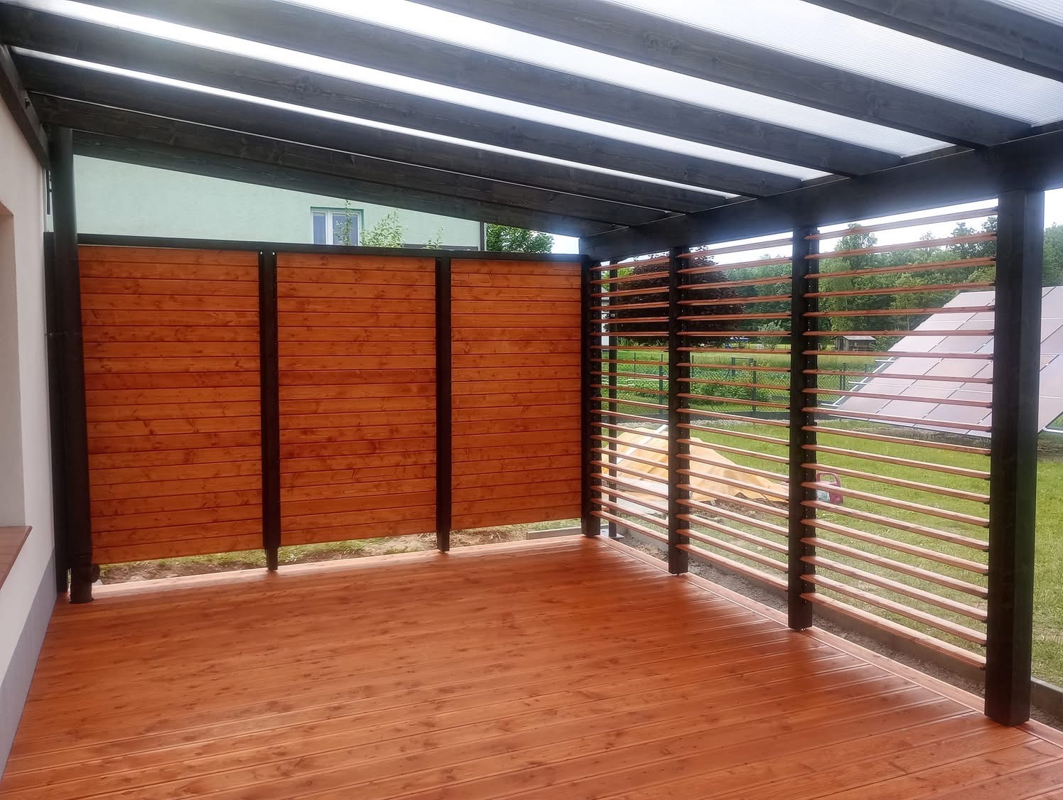 Zadaszony taras z drewnianą podłogą i ażurowymi ścianami, z widokiem na zielony ogród i panele słoneczne w tle. Ciemne belki konstrukcyjne.