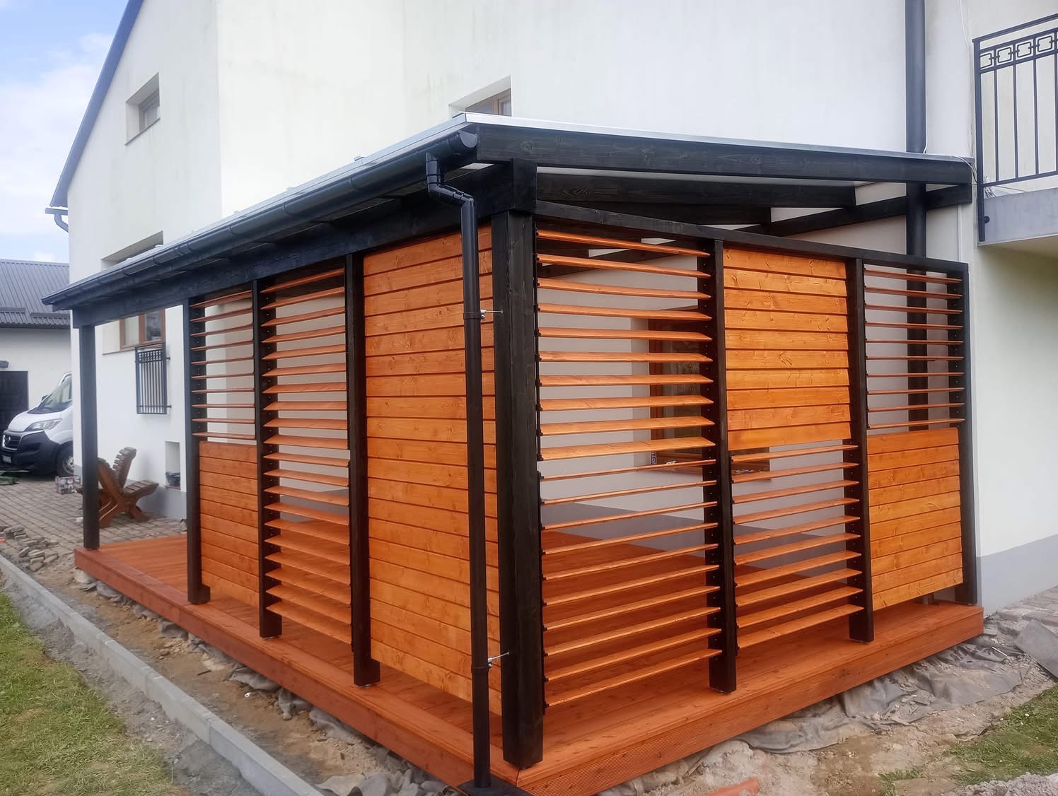 Nowoczesna pergola z drewnianymi lamelami w kolorze miodowym, z czarną konstrukcją, przylegająca do białego budynku. Widoczny fragment tarasu i otoczenie domu.