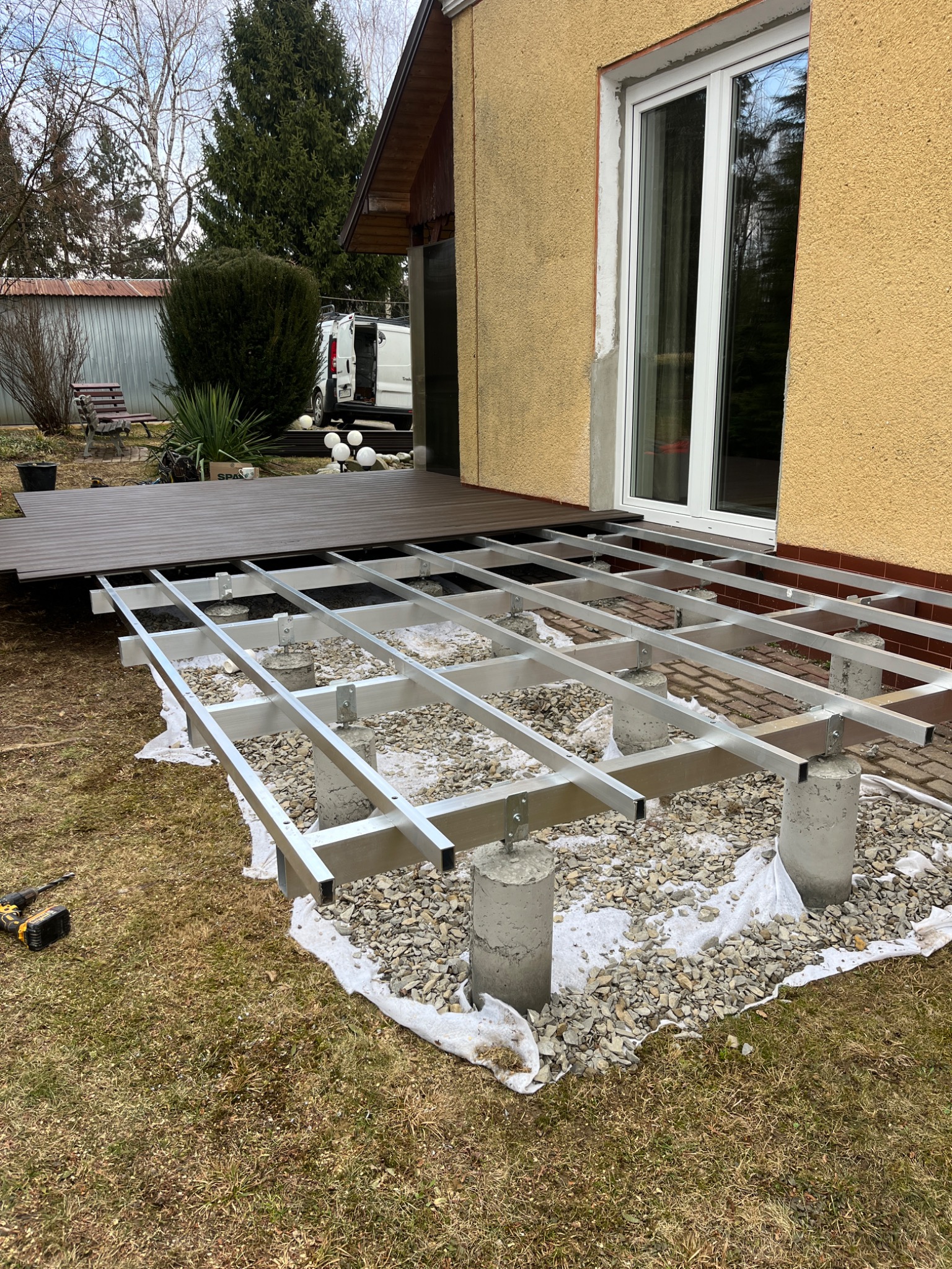 Montaż tarasu kompozytowego: aluminiowa konstrukcja na betonowych słupkach, częściowo ułożone deski. Widoczny wkrętak akumulatorowy i dom z oknem.