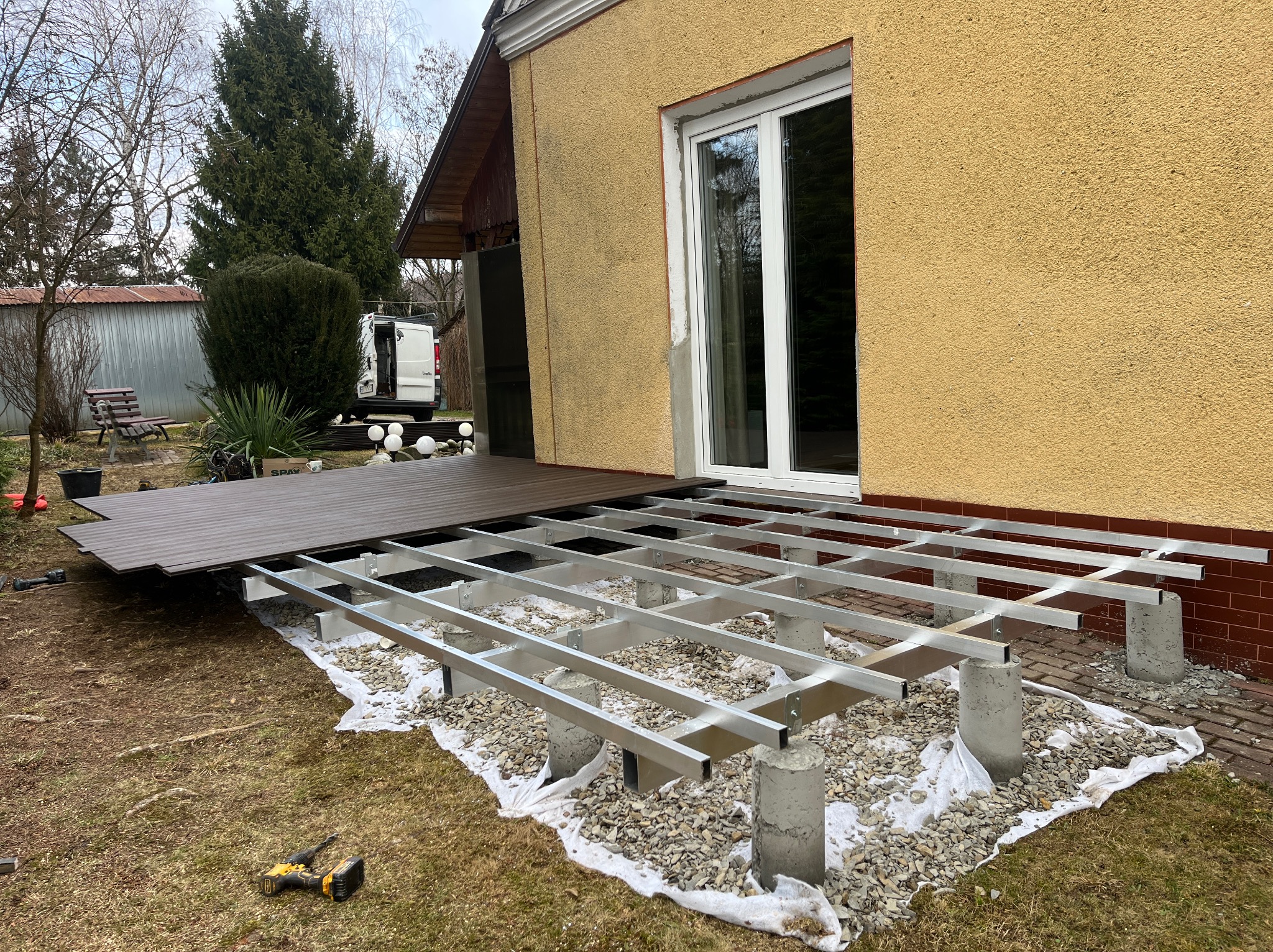 Montaż tarasu kompozytowego: aluminiowa konstrukcja na betonowych słupkach, częściowo ułożone deski w odcieniach brązu, narzędzia obok. Widok na budynek z oknem.