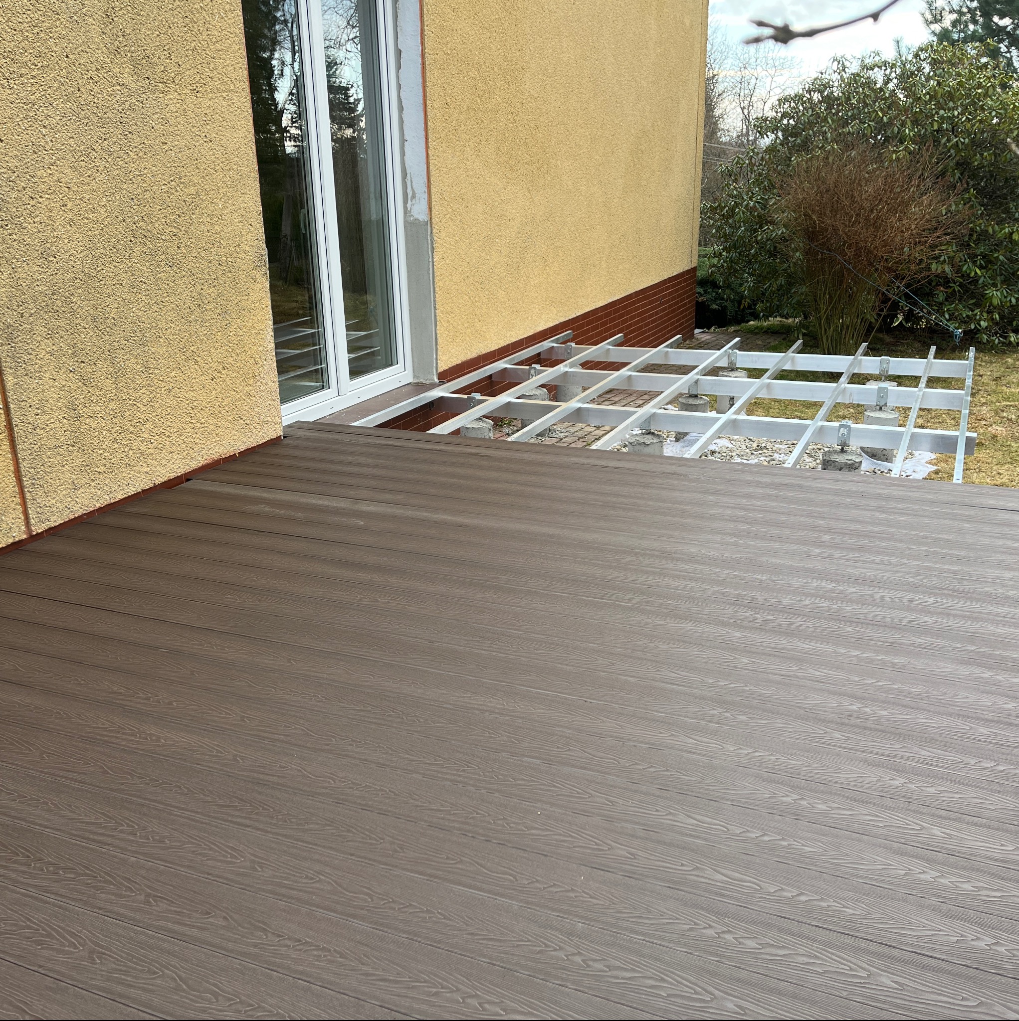 Montaż tarasu kompozytowego: widok na częściowo ułożone deski w odcieniach brązu i aluminiową konstrukcję nośną na betonowych podstawach przy ścianie budynku.
