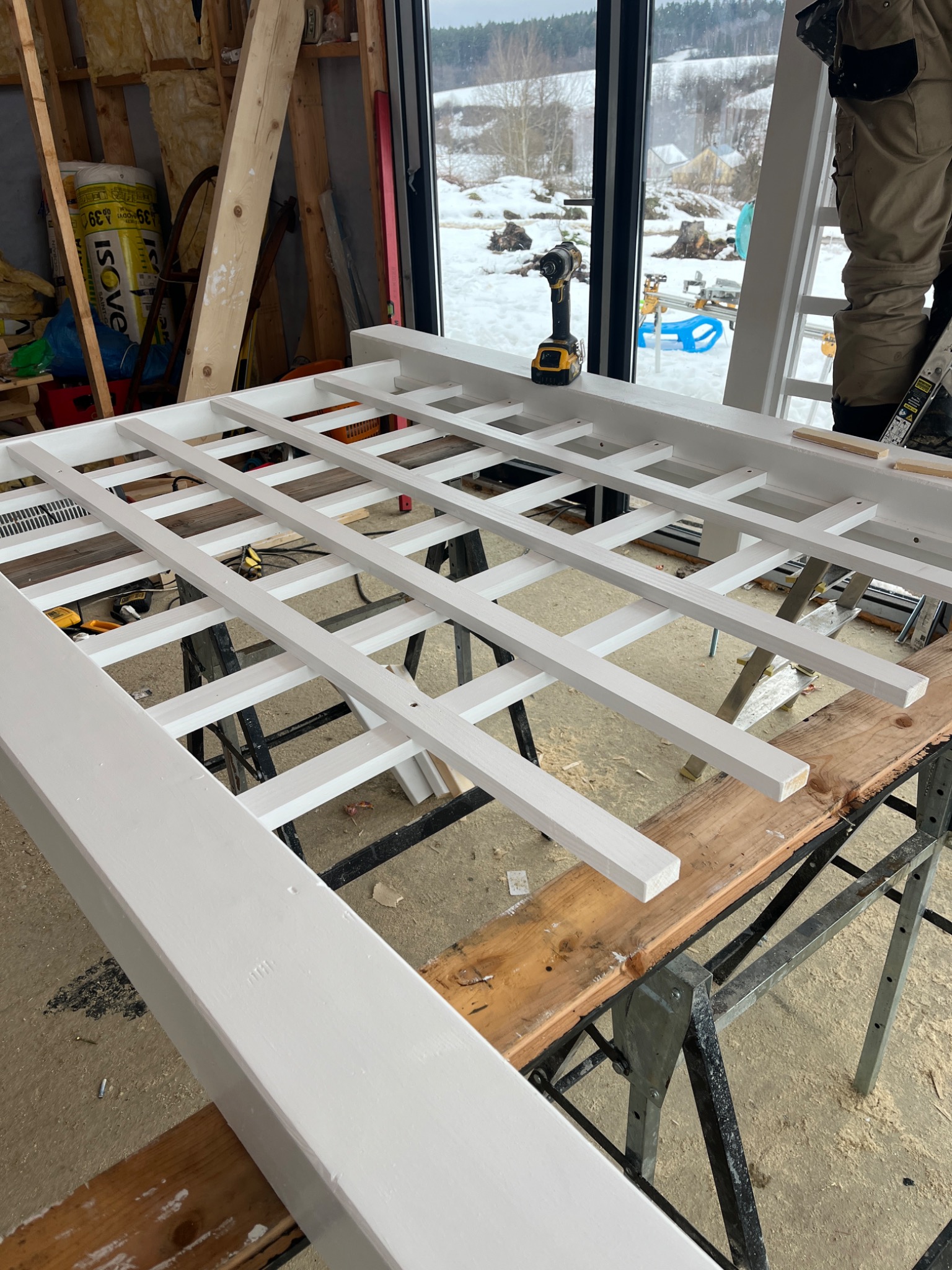 Biała, drewniana konstrukcja kratowa na stole warsztatowym, w tle okno z zimowym krajobrazem. Widać wkrętarkę i fragment osoby pracującej nad konstrukcją.
