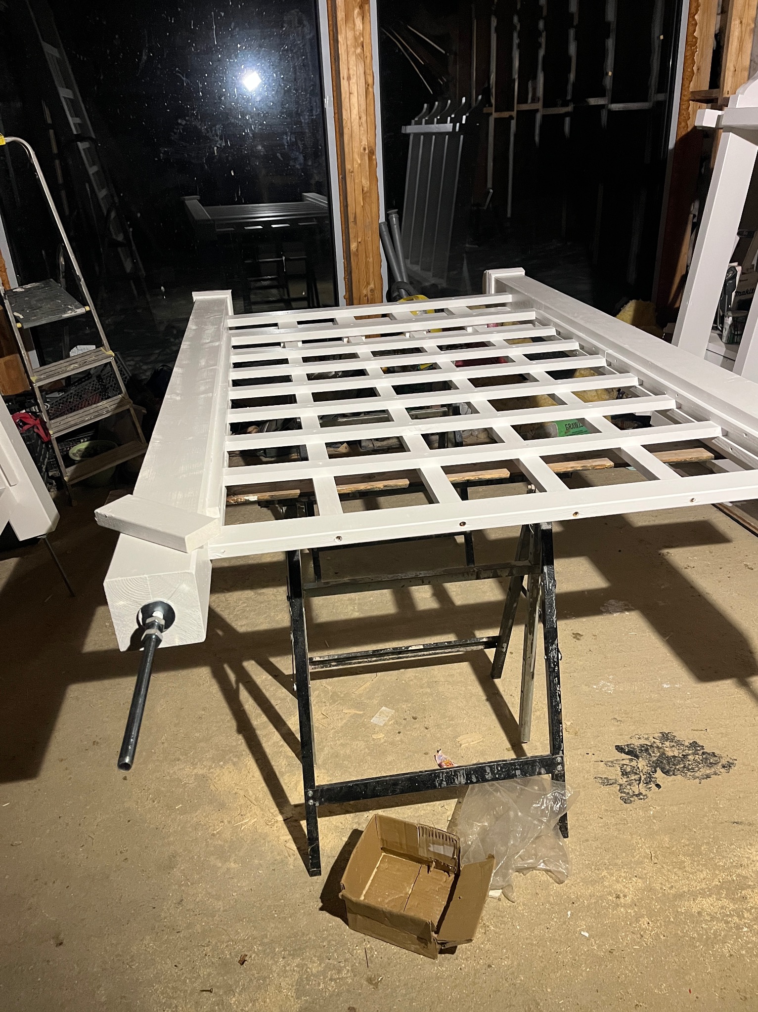 Biała, drewniana konstrukcja kratowa na czarnym stojaku warsztatowym, z widocznym zaciskiem stolarskim. W tle okno i drabina.