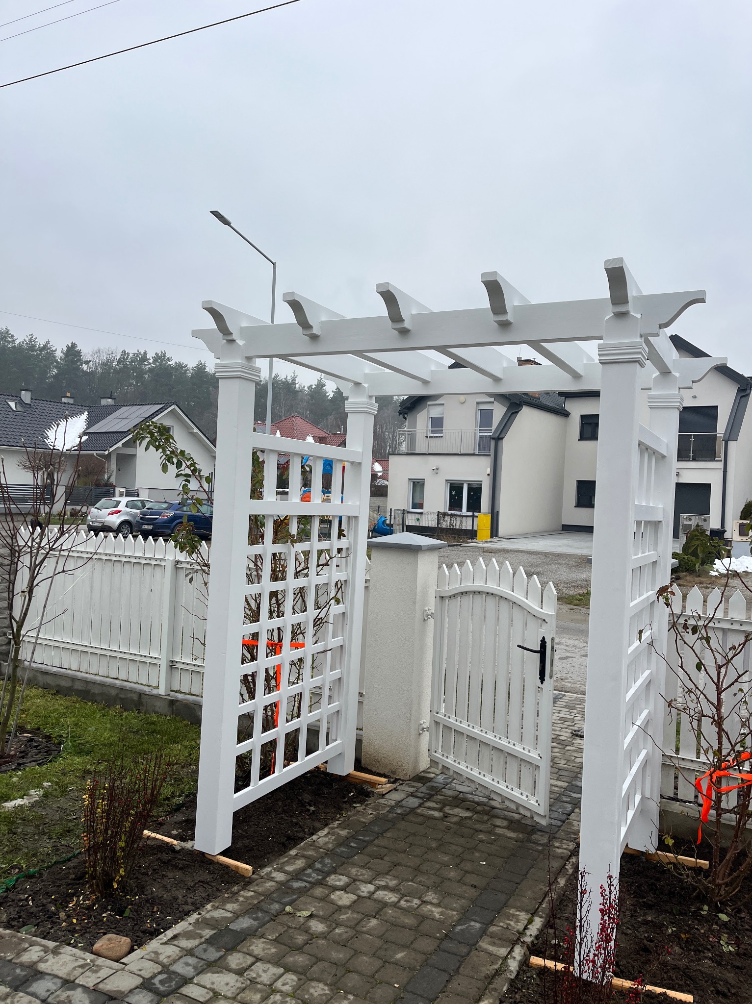 Biała pergola z kratką i otwartą furtką, prowadząca do ogrodu. W tle domy i szara, pochmurna pogoda. Widoczne detale wykończenia pergoli i kostka brukowa.