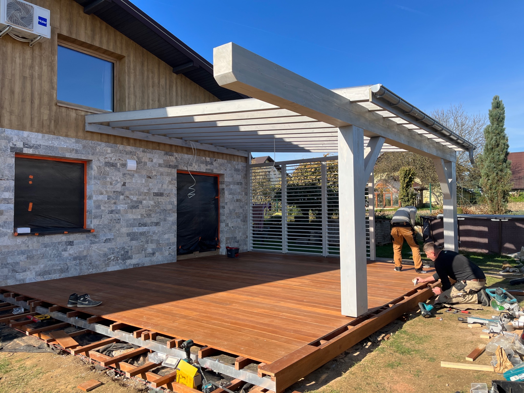 Montaż drewnianego tarasu z pergolą przy domu. Widoczne narzędzia, deski i pracownicy podczas instalacji. Jasne, słoneczne oświetlenie.