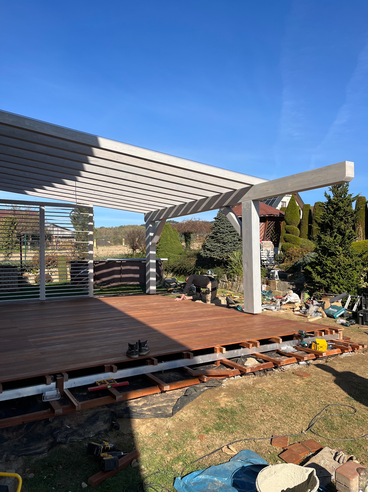 Montaż tarasu drewnianego z pergolą w ogrodzie. Widoczne narzędzia i elementy konstrukcyjne. Częściowo ukończona podłoga z desek. Słoneczny dzień.