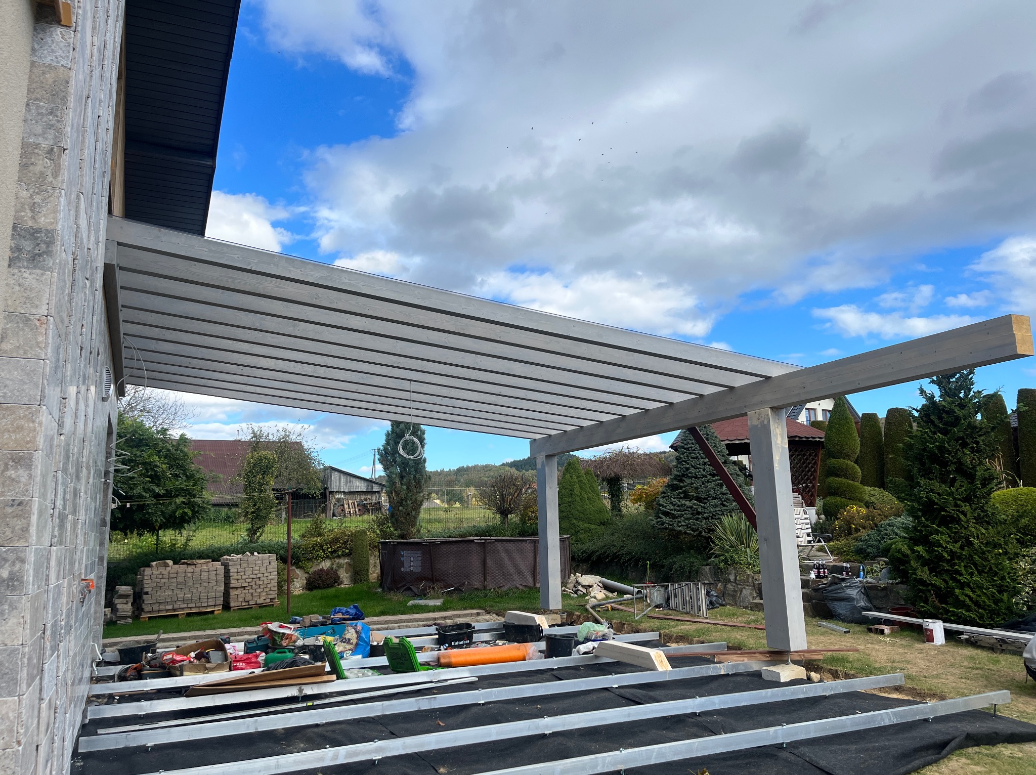 Montaż zadaszenia tarasu z szarych belek na czarnej membranie. Widoczne narzędzia i materiały budowlane. W tle dom i ogród w słoneczny dzień.