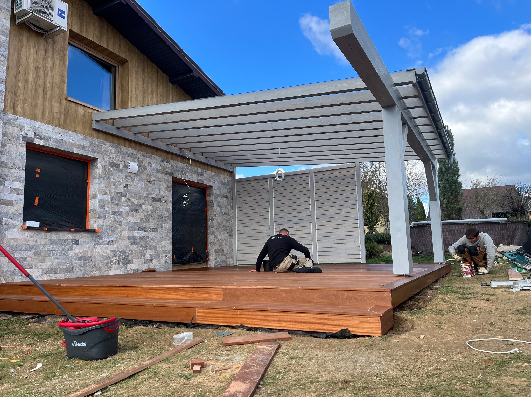 Montaż drewnianego tarasu z szarą pergolą przy domu z elewacją z kamienia i drewna. Dwóch monterów pracuje nad wykończeniem tarasu. Widoczny kubeł Vileda.