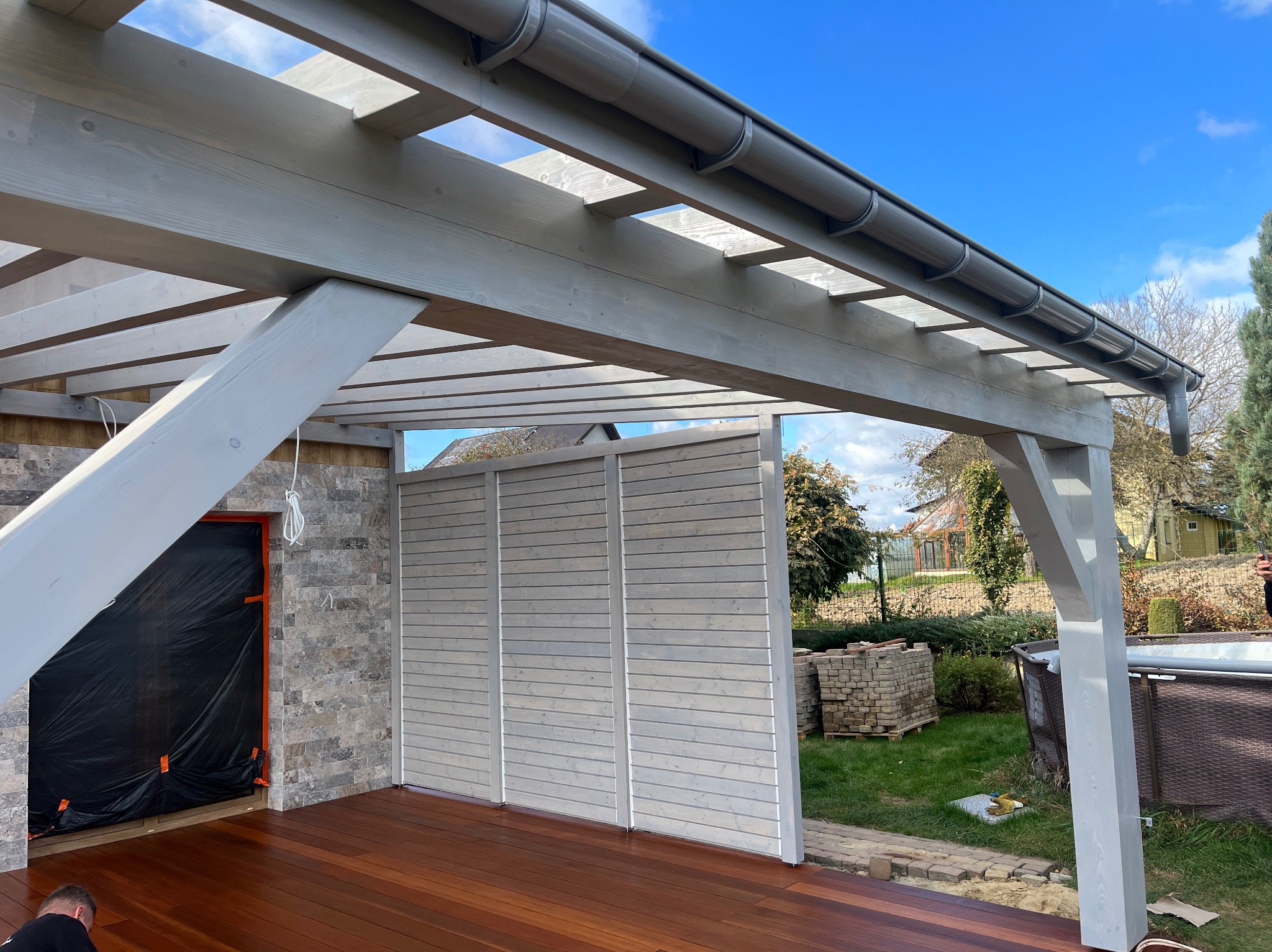 Zadaszona, drewniana konstrukcja z szarymi elementami i brązową podłogą. Widoczne deski, ściana z paneli i rynna. W tle zieleń ogrodu i błękitne niebo.