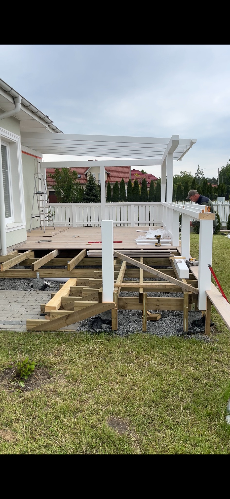 Budowa tarasu drewnianego z białą pergolą. Widoczna konstrukcja nośna, częściowo ułożona deska tarasowa i pracownik montujący balustradę. W tle dom jednorodzinny.