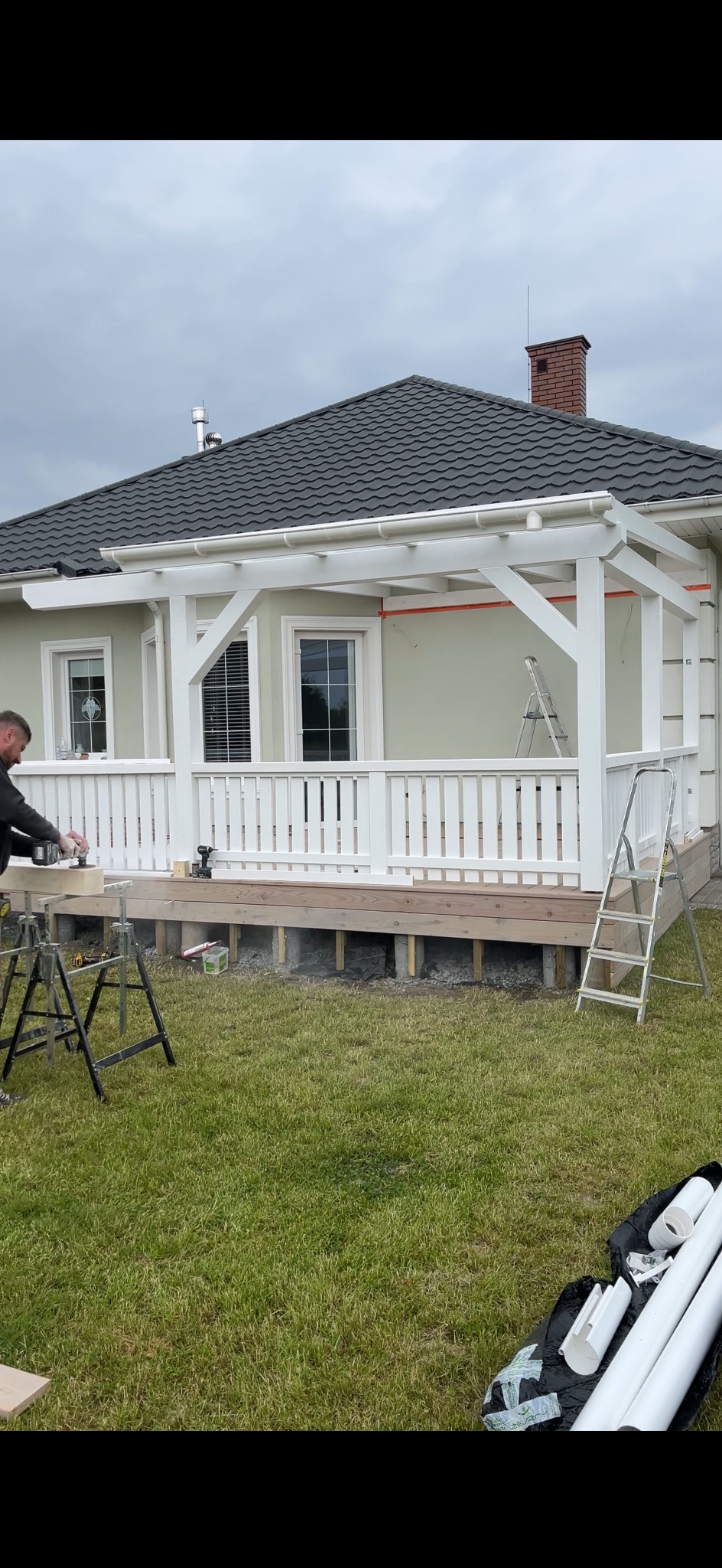 Budowa białej pergoli przy domu. Widoczny pracownik szlifujący drewno, narzędzia i części konstrukcji na trawniku. Etap montażu tarasu z balustradą.