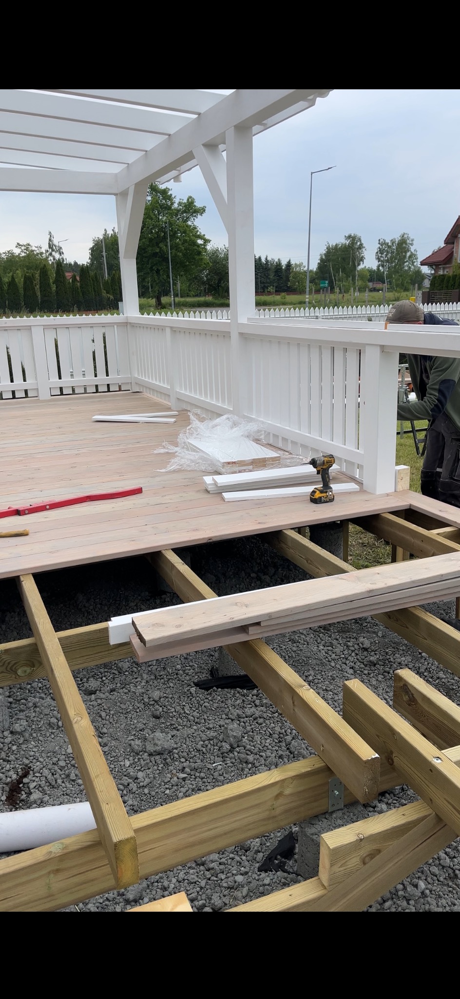 Budowa tarasu: drewniana konstrukcja szkieletowa z częściowo ułożonymi deskami, biała balustrada i pergola w tle. Widoczne narzędzia i materiały budowlane.