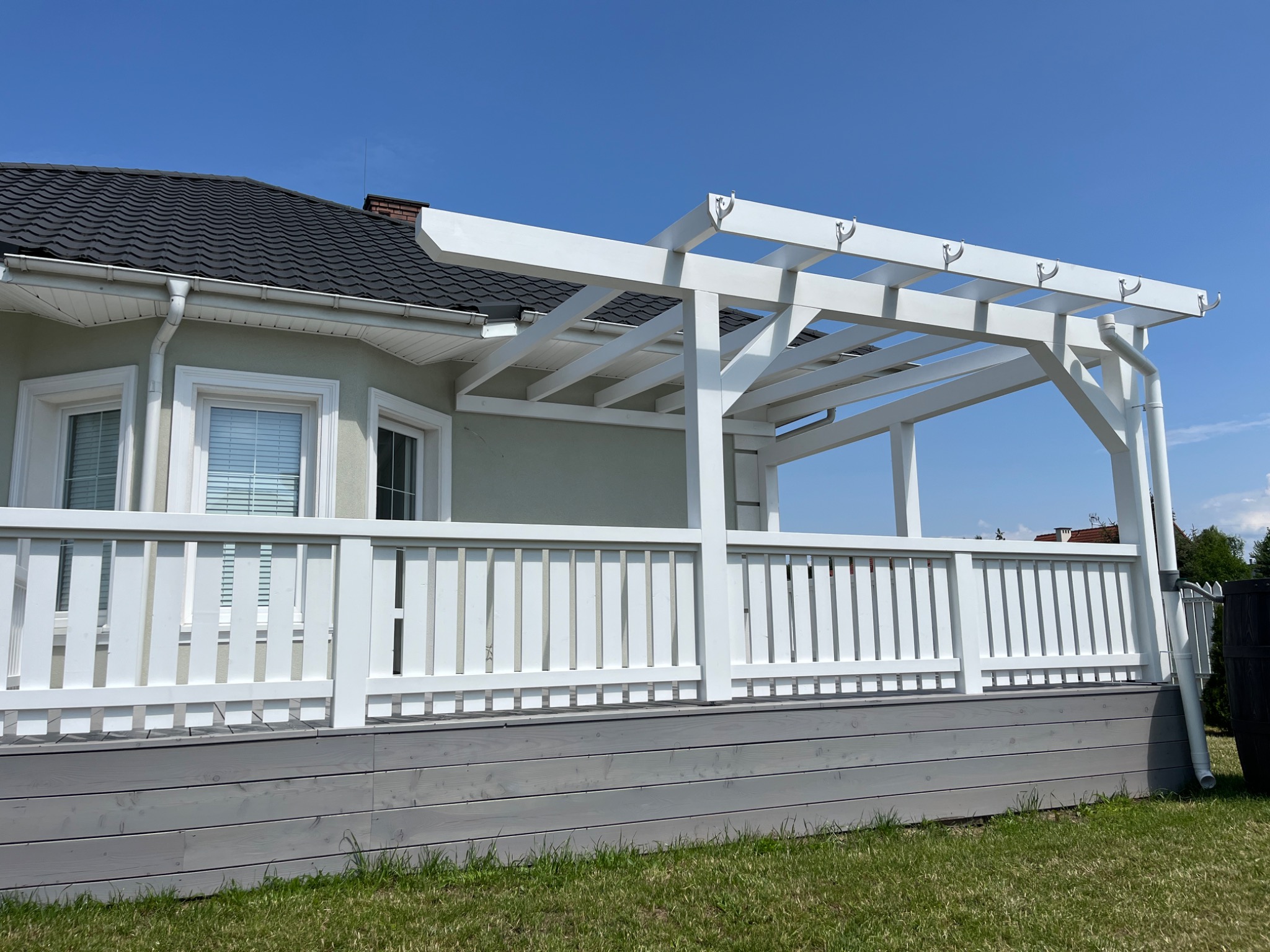 Biała pergola z balustradą na szarym tarasie, przylegająca do domu z ciemnym dachem. Elementy konstrukcyjne z hakami. Jasne, słoneczne ujęcie.
