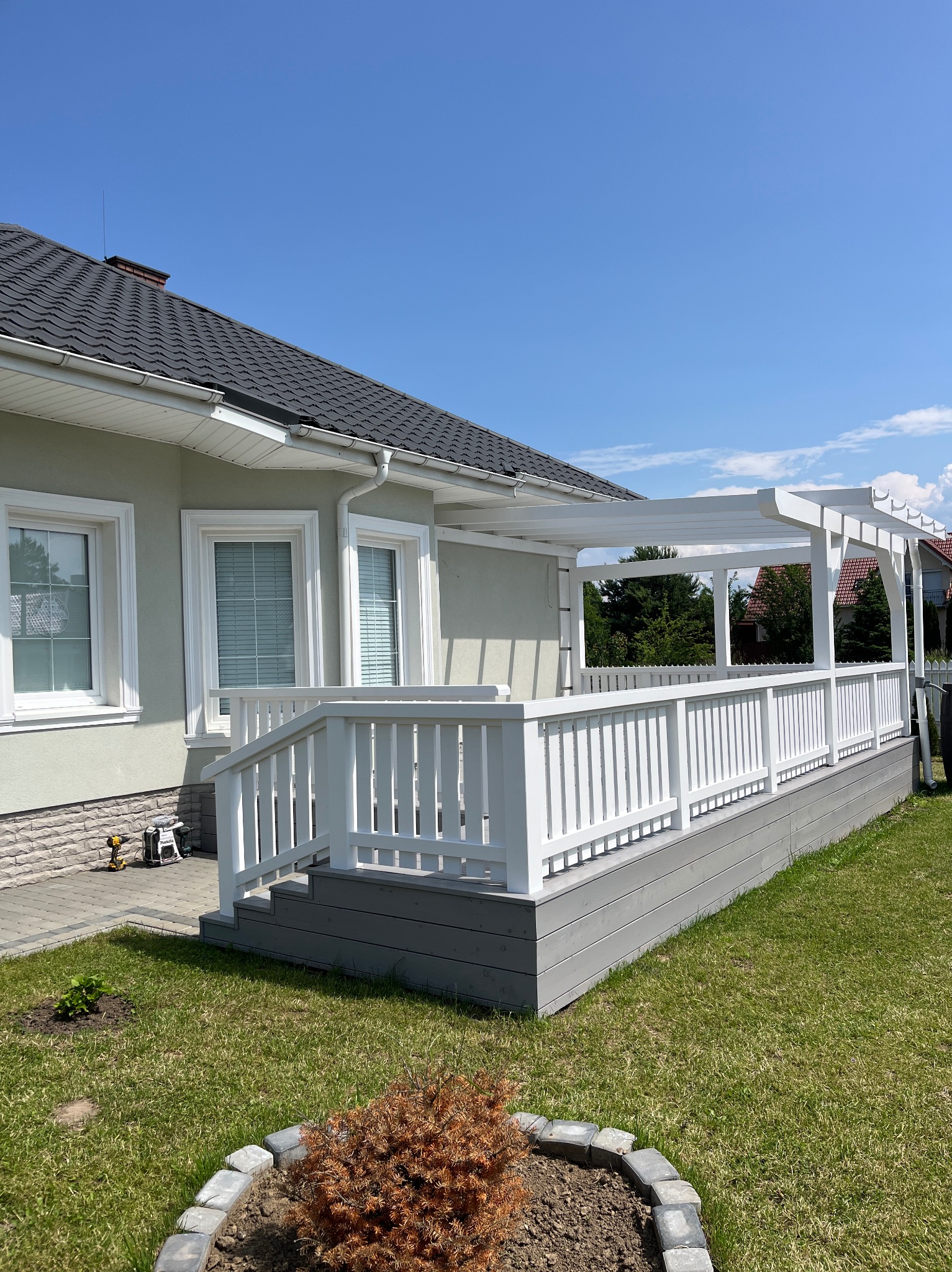 Biała pergola z balustradą na szarym tarasie przylegającym do domu. Widoczne narzędzia budowlane. Błękitne niebo w tle. Słoneczny dzień, trawnik.