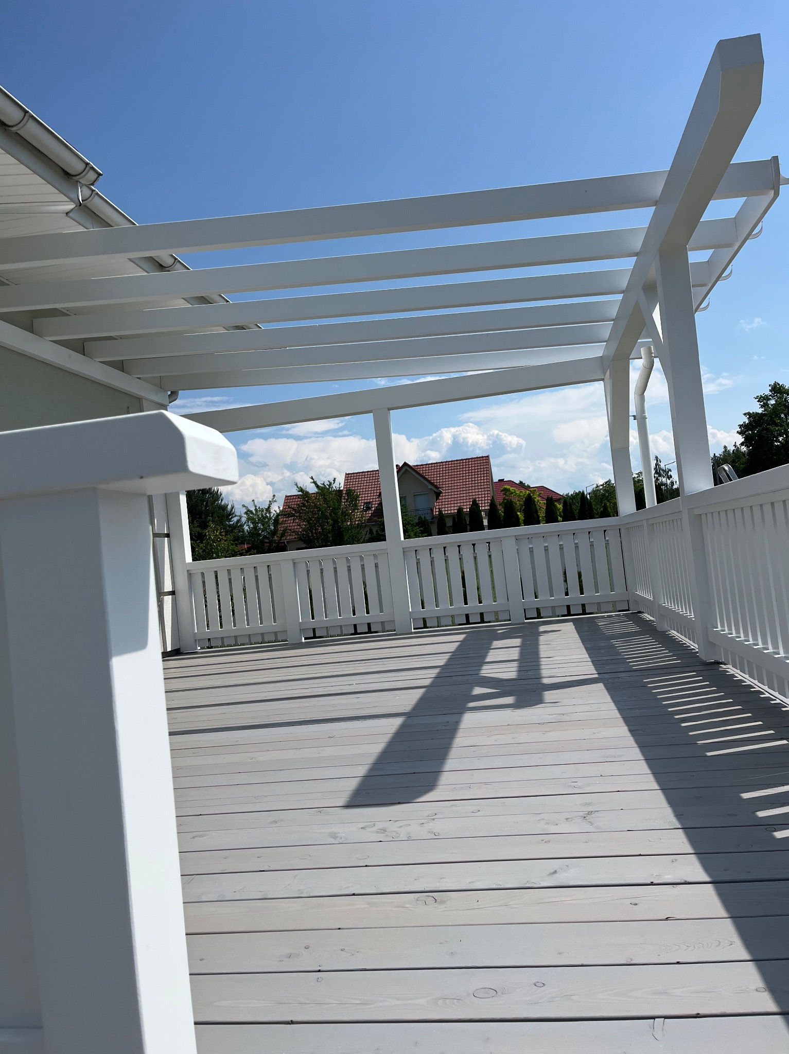 Biała pergola z drewnianym tarasem w słoneczny dzień. Widok na dom z czerwonym dachem przez białe balustrady. Cienie tworzą geometryczne wzory na podłodze.