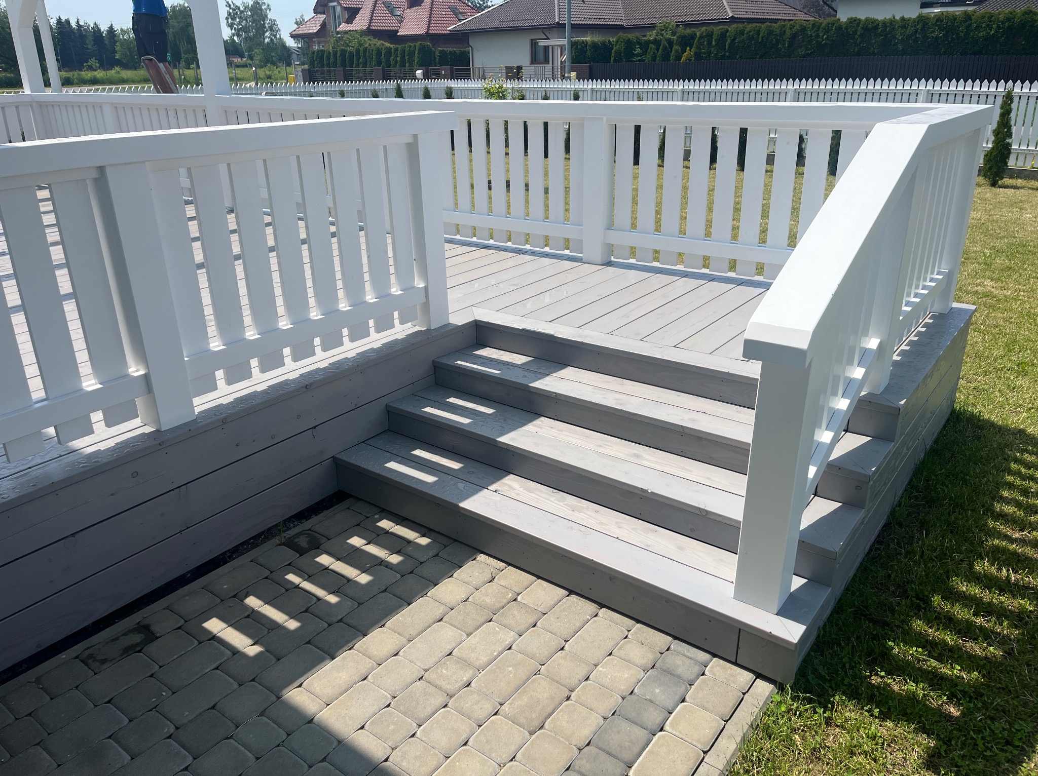 Nowoczesny taras z szarej deski i białymi balustradami, schody z kostki brukowej, w tle dom z czerwonym dachem i zielony trawnik. Jasne, słoneczne oświetlenie.