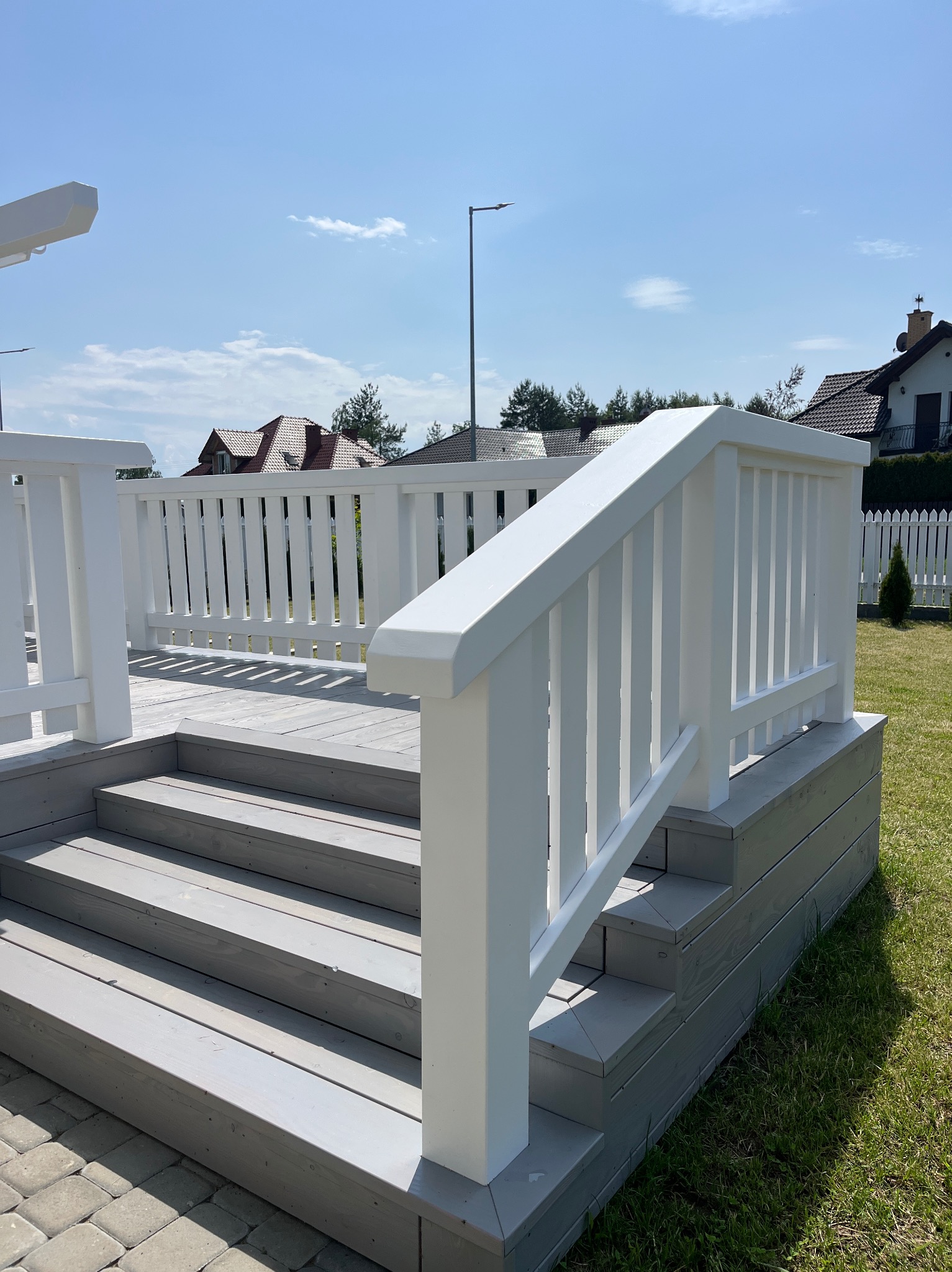 Jasnoszary taras z białymi balustradami i schodami, widok z dołu, w tle domy i błękitne niebo. Estetyczne wykończenie, schludny design.