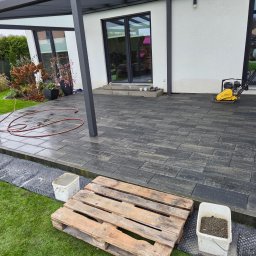 Solidbruk - Nowoczesny taras z ciemnej kostki, z widoczną ubijarką i elementami przygotowawczymi. Paleta i wiadro na trawie sugerują trwające prace wykończeniowe.