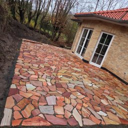 Solidbruk - Ułożona ścieżka z nieregularnych płyt piaskowca w odcieniach czerwieni i beżu, prowadząca do jasnego, ceglanego domu z białymi oknami. Tło stanowią drzewa.