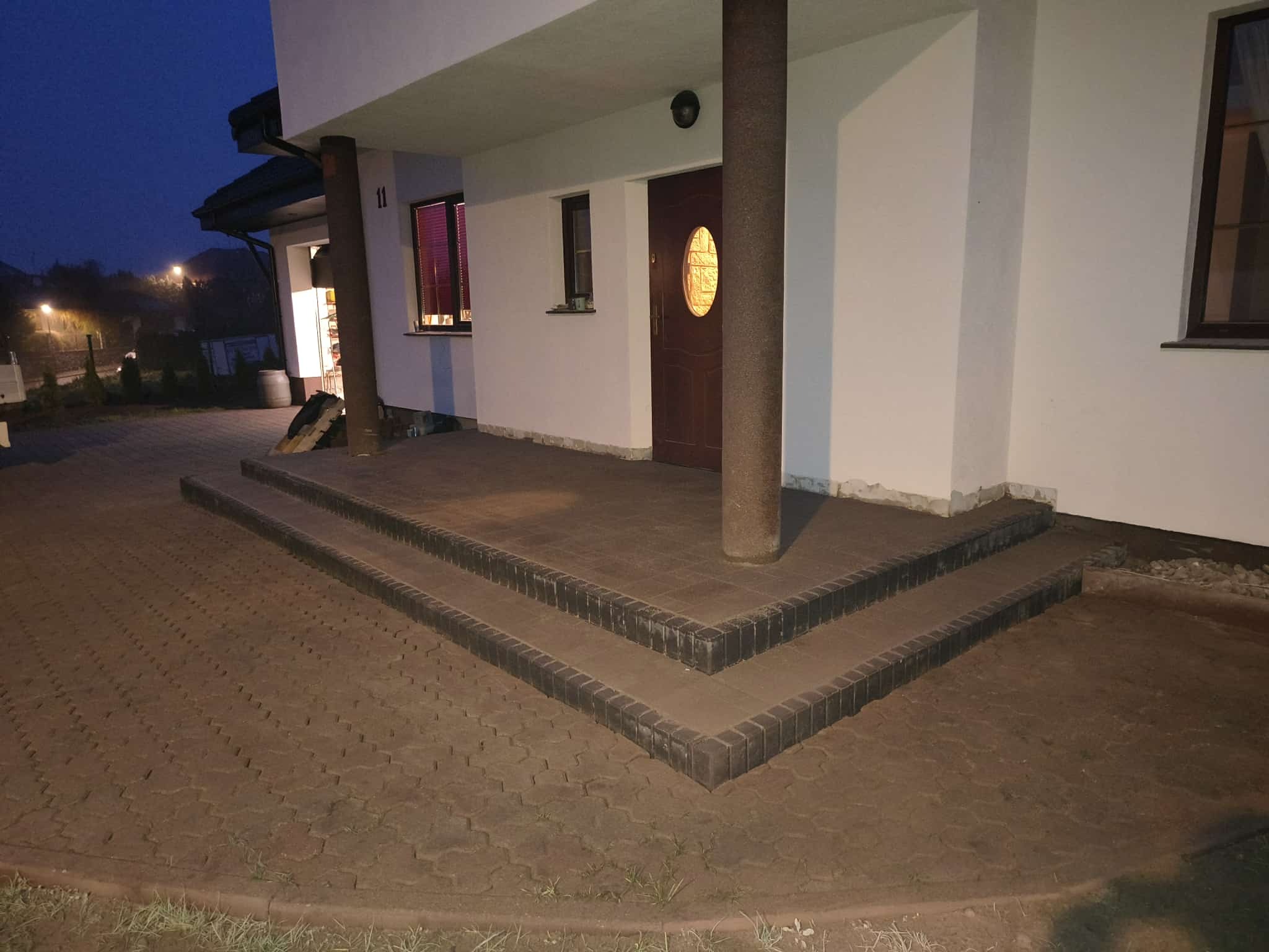 Ułożona kostka brukowa przed domem z trzema stopniami i kolumną. Ciemna kolorystyka kostki, widoczne oświetlenie zewnętrzne. Ujęcie z dołu, wieczorne.