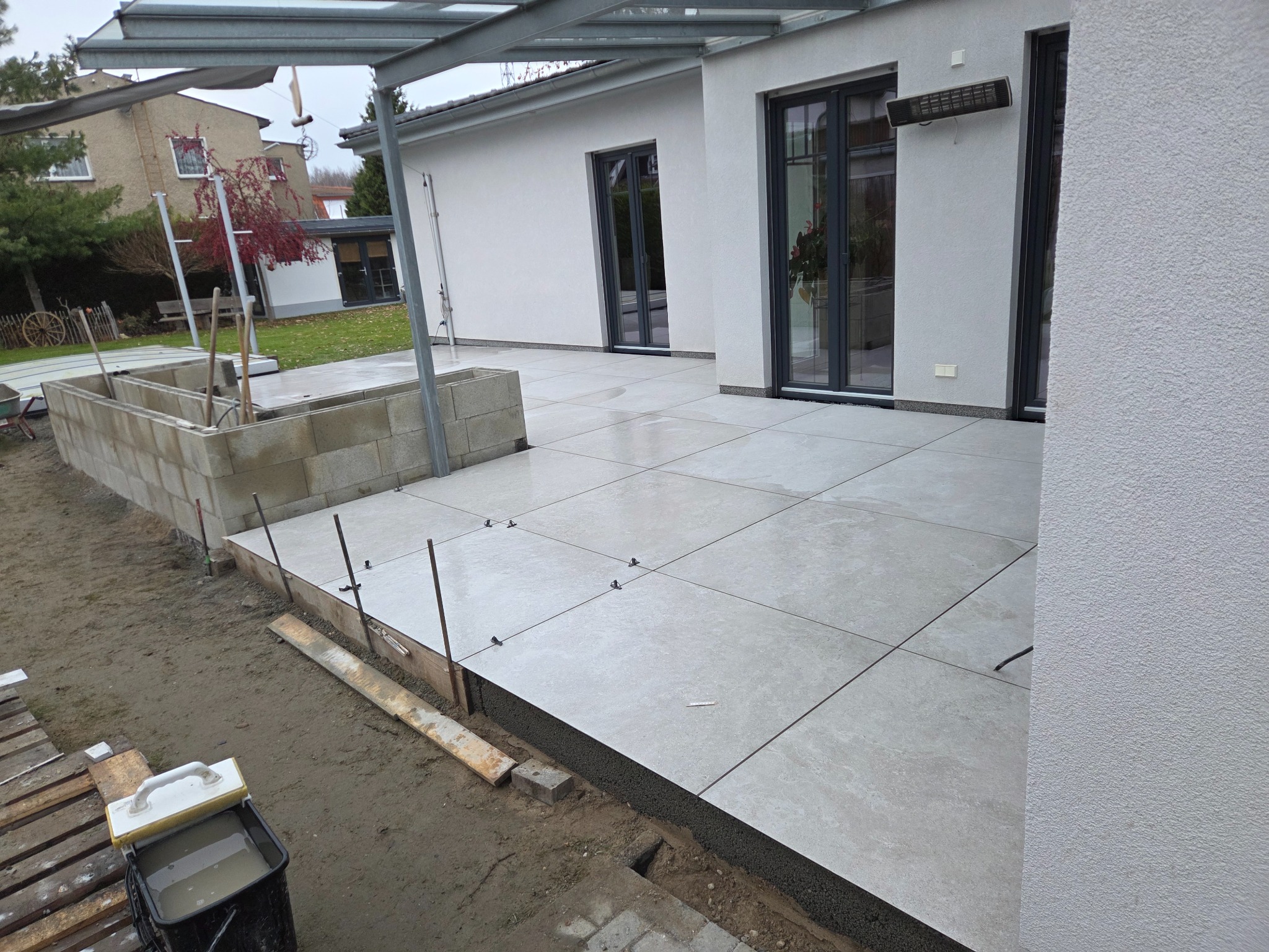 Jasnoszara, kwadratowa nawierzchnia tarasu w trakcie układania, z widocznymi elementami mocującymi i narzędziami. Tło stanowi biały budynek z szarymi oknami.