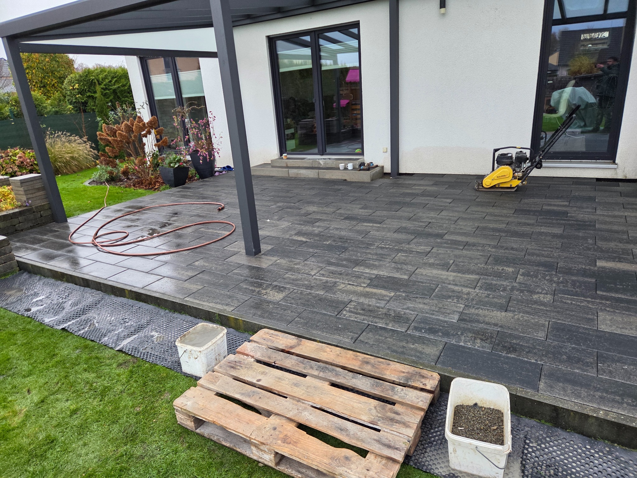 Nowoczesny taras z ciemnej kostki, z widoczną ubijarką i elementami przygotowawczymi. Paleta i wiadro na trawie sugerują trwające prace wykończeniowe.