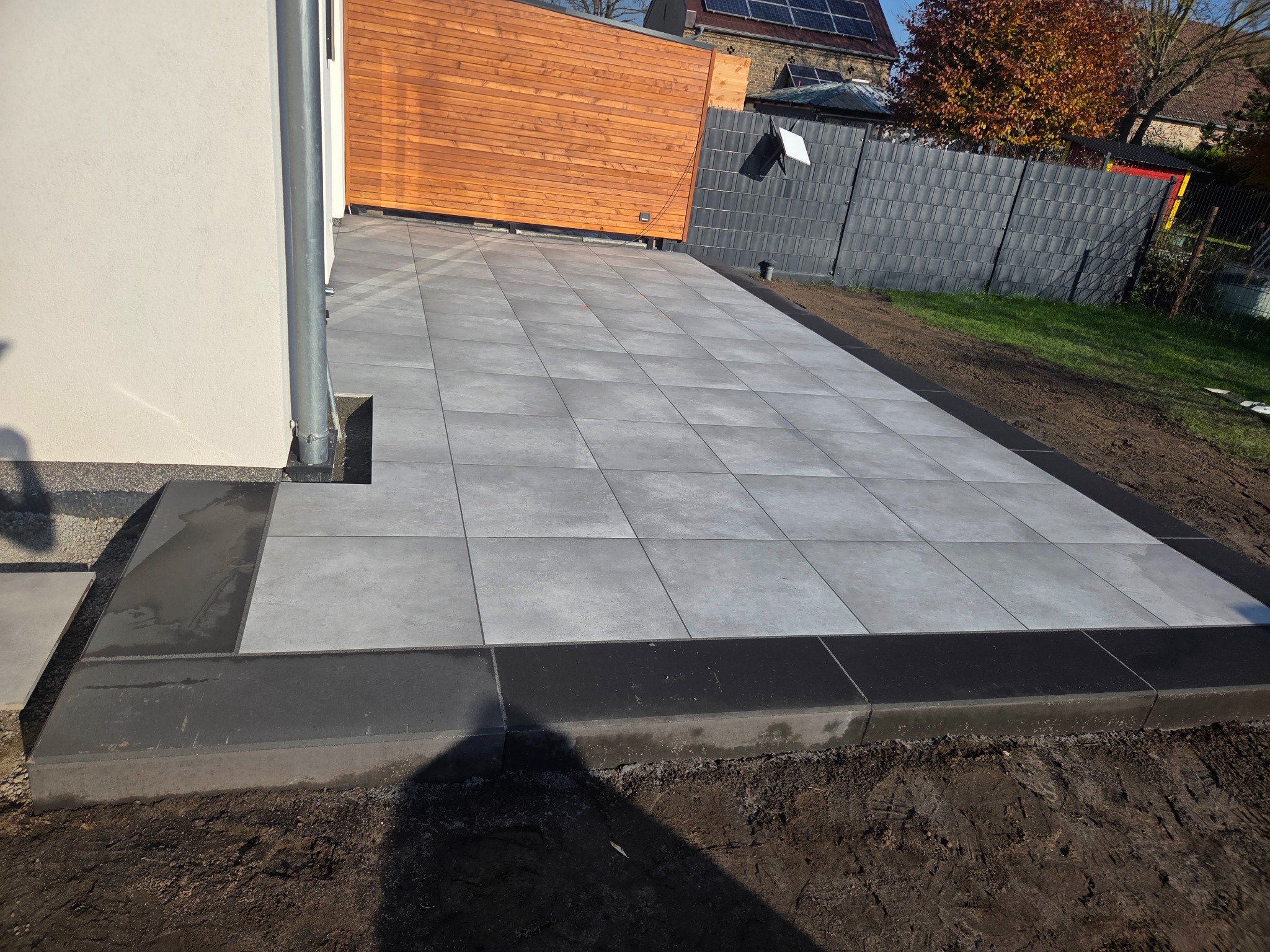 Nowoczesny taras z szarych płyt, obrzeża z ciemnego kamienia. W tle drewniana brama i szary płot panelowy. Widoczne nasadzenia i fragment budynku z rynną.