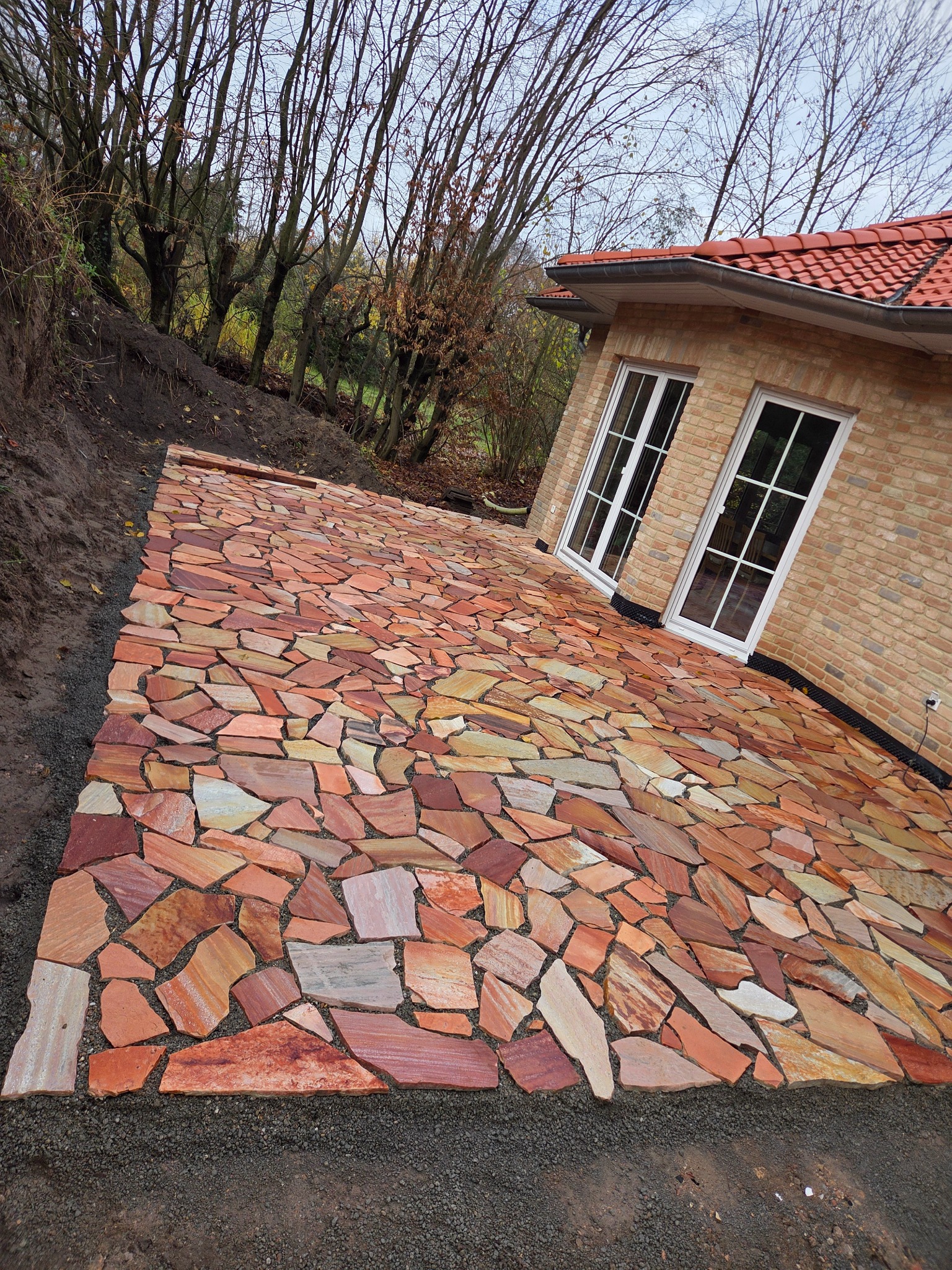 Ułożona ścieżka z nieregularnych płyt piaskowca w odcieniach czerwieni i beżu, prowadząca do jasnego, ceglanego domu z białymi oknami. Tło stanowią drzewa.