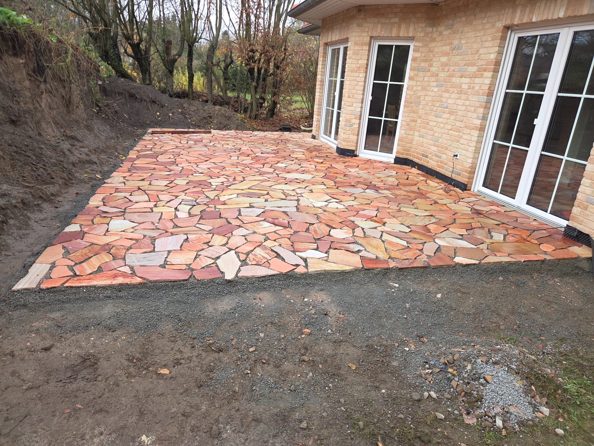 Wykończony taras z nieregularnych, czerwono-beżowych płyt kamiennych przy budynku z cegły. Widoczne białe ramy okienne i przygotowane podłoże.