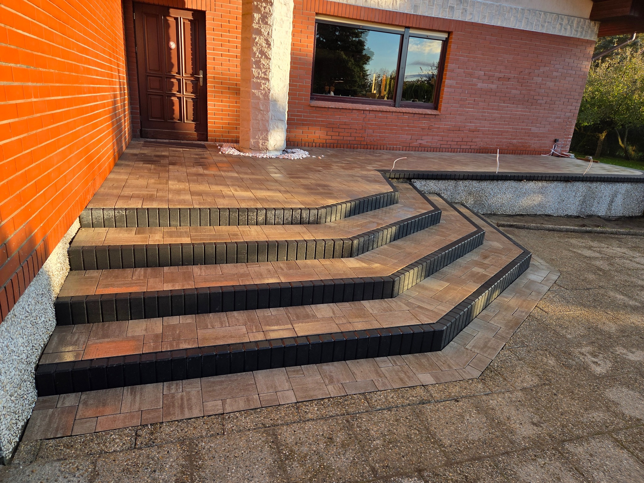 Schody wejściowe z kostki brukowej w odcieniach brązu i czerni, prowadzące do domu z cegły. Widoczne detale wykończenia i estetyczne połączenie z elewacją.