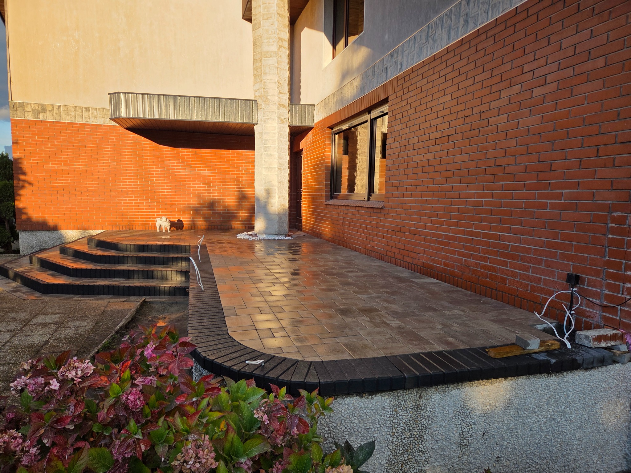 Błyszcząca, brązowa kostka brukowa na tarasie domu z ceglaną ścianą, obrzeżem z ciemnej kostki i kwiatami w pierwszym planie. Widoczny fragment schodów i kolumna.