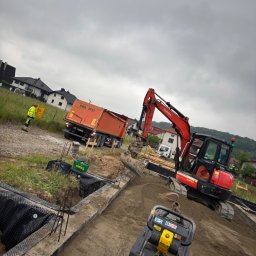 Firma Handlowo Transportowa Żyła Roman - Prace ziemne: czerwona mini koparka Kubota, zagęszczarka i pomarańczowa wywrotka na placu budowy. Widoczny pracownik w odblaskowej kamizelce. Etap przygotowania podłoża.