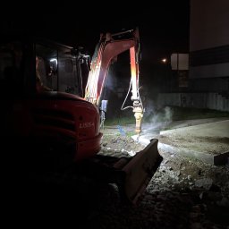 Firma Handlowo Transportowa Żyła Roman - Czerwona koparka Kubota U55-4 z włączonymi światłami pracuje nocą, rozbijając betonowy fundament. Widoczne gruz i pył. Praca w toku.