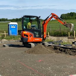 Firma Handlowo Transportowa Żyła Roman - Pomarańczowa mini koparka Kubota KX018-4 w trakcie prac ziemnych na budowie, z widocznym operatorem i przenośną toaletą w tle.
