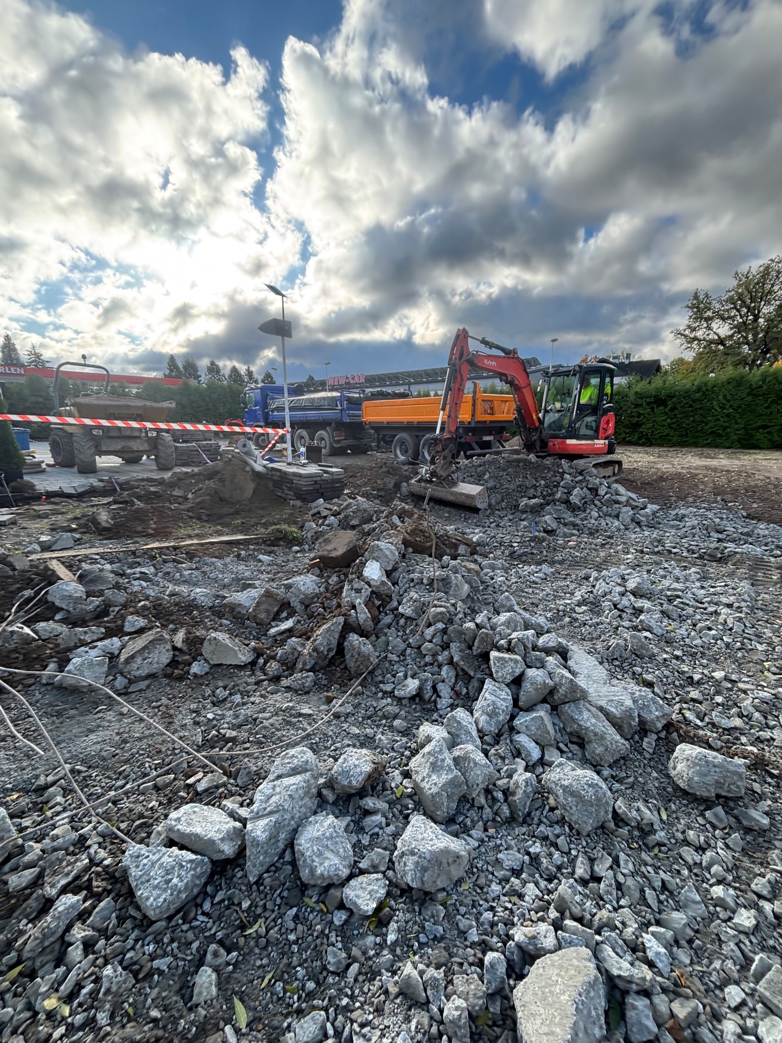 Prace ziemne w Frydrychowicach: Mini koparka Kubota na placu budowy, gruz i kamienie w pierwszym planie, w tle ciężarówki i maszyny budowlane pod pochmurnym niebem.