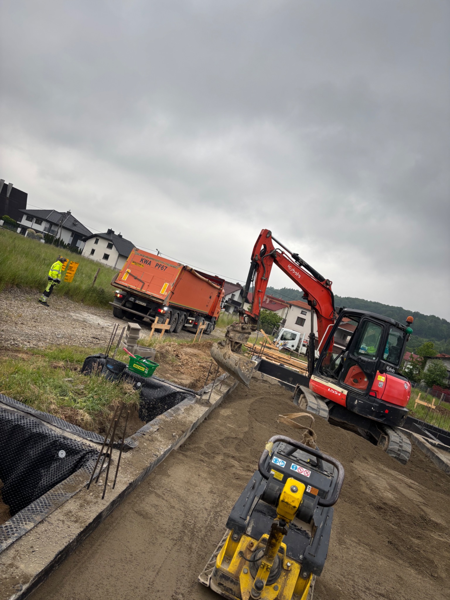 Prace ziemne: czerwona mini koparka Kubota, zagęszczarka i pomarańczowa wywrotka na placu budowy. Widoczny pracownik w odblaskowej kamizelce. Etap przygotowania podłoża.